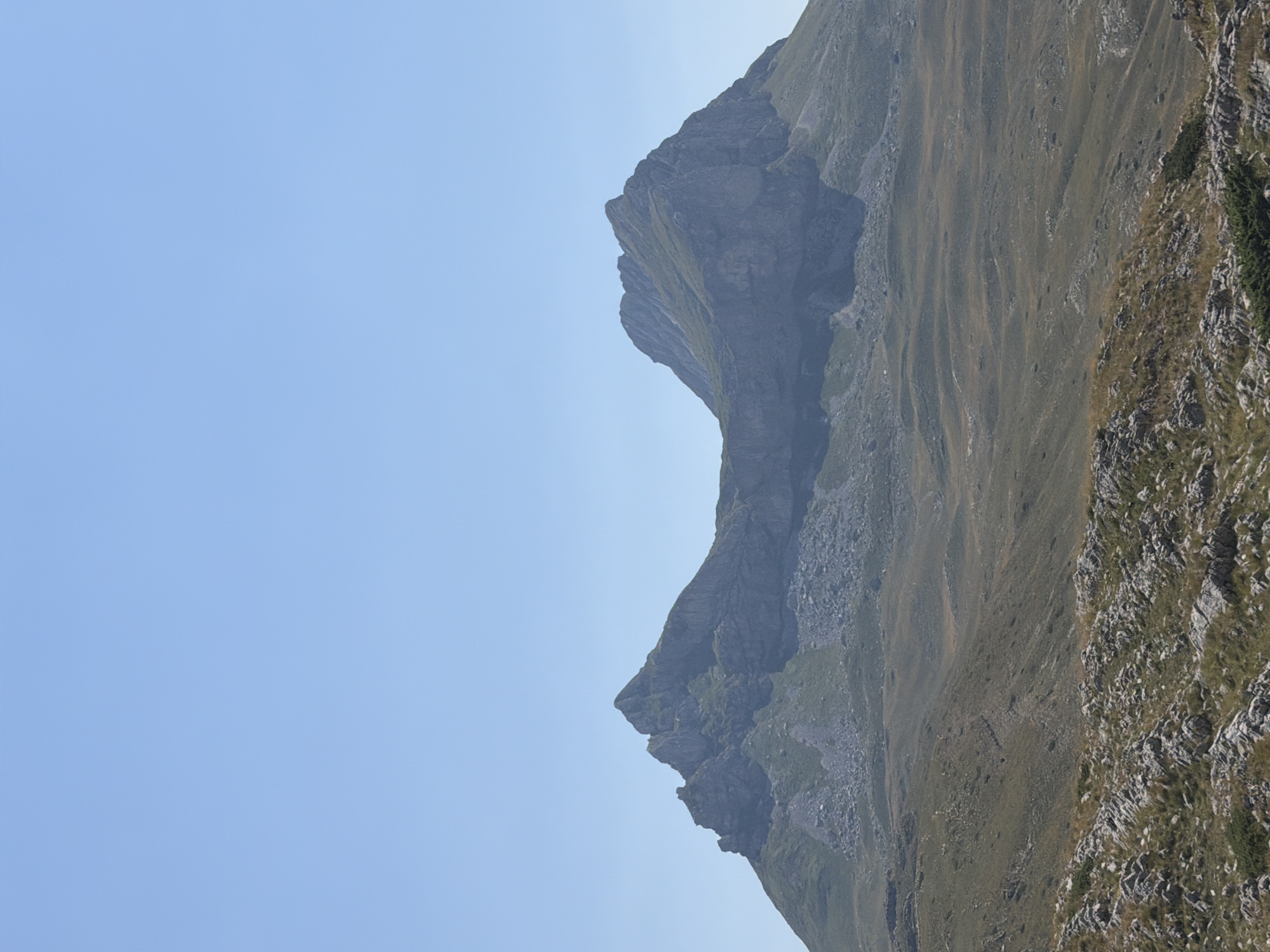 The image depicts a wide, sweeping landscape dominated by rugged mountains under a clear blue sky. In the foreground, the terrain is rocky with patches of dry grass. Further back, rolling hills covered in sparse vegetation lead up to the base of two prominent, rocky mountain peaks. The larger, left peak has a jagged, sharp summit, while the right peak is broader and more rounded, with steep, scree-covered slopes. The overall environment suggests a mountainous, possibly arid region, with no signs of human habitation or activity visible in the scene.