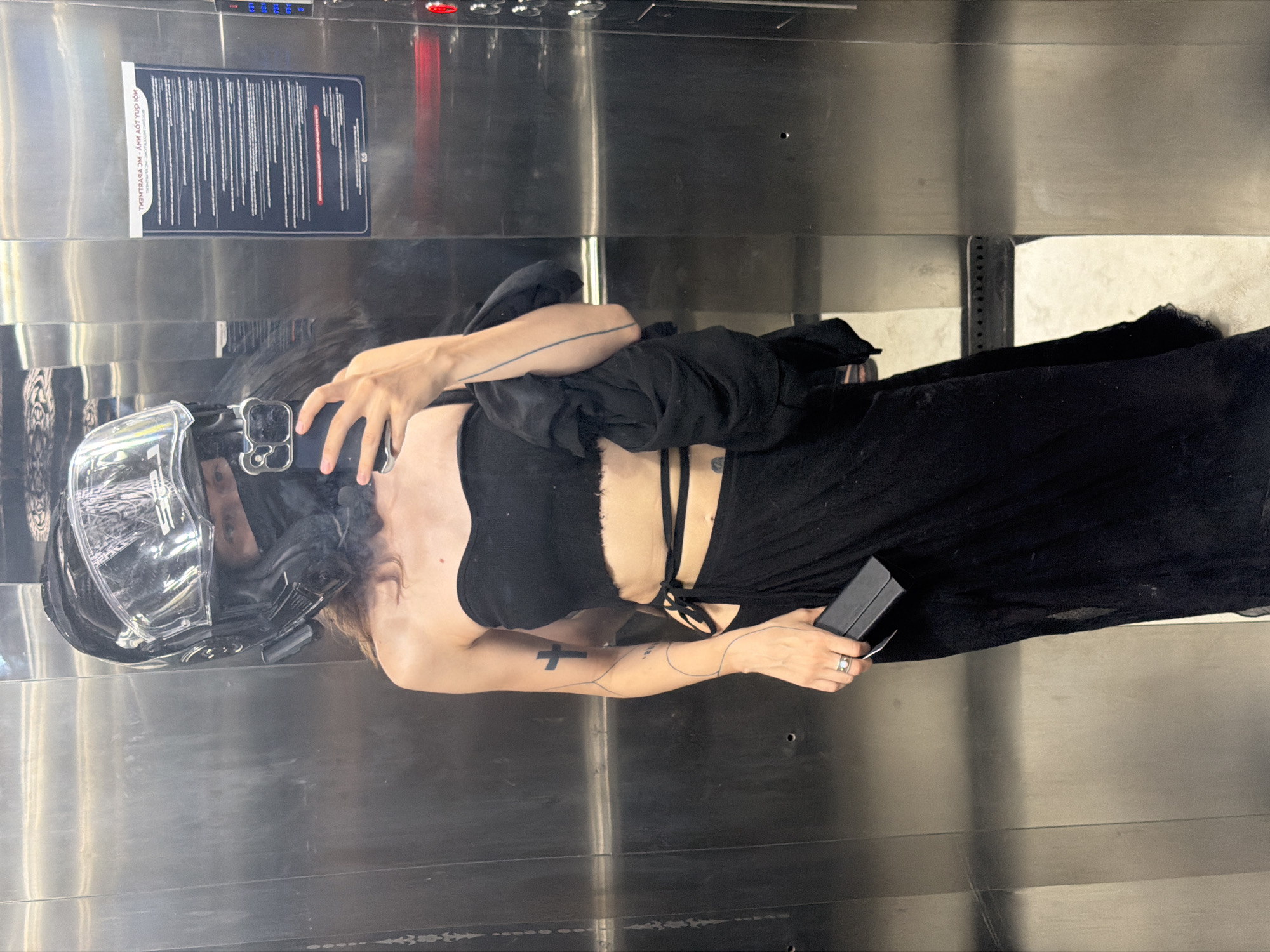 A person wearing a motorcycle helmet and black clothing stands in an elevator with stainless steel walls. The person is holding a smartphone in one hand and a small black rectangular object in the other. A sign is affixed to the elevator wall. The person is facing the elevator wall and taking a selfie or photograph in the reflection of the stainless steel. There is no evidence indicating that this image was taken in Đà Nẵng, Vietnam.