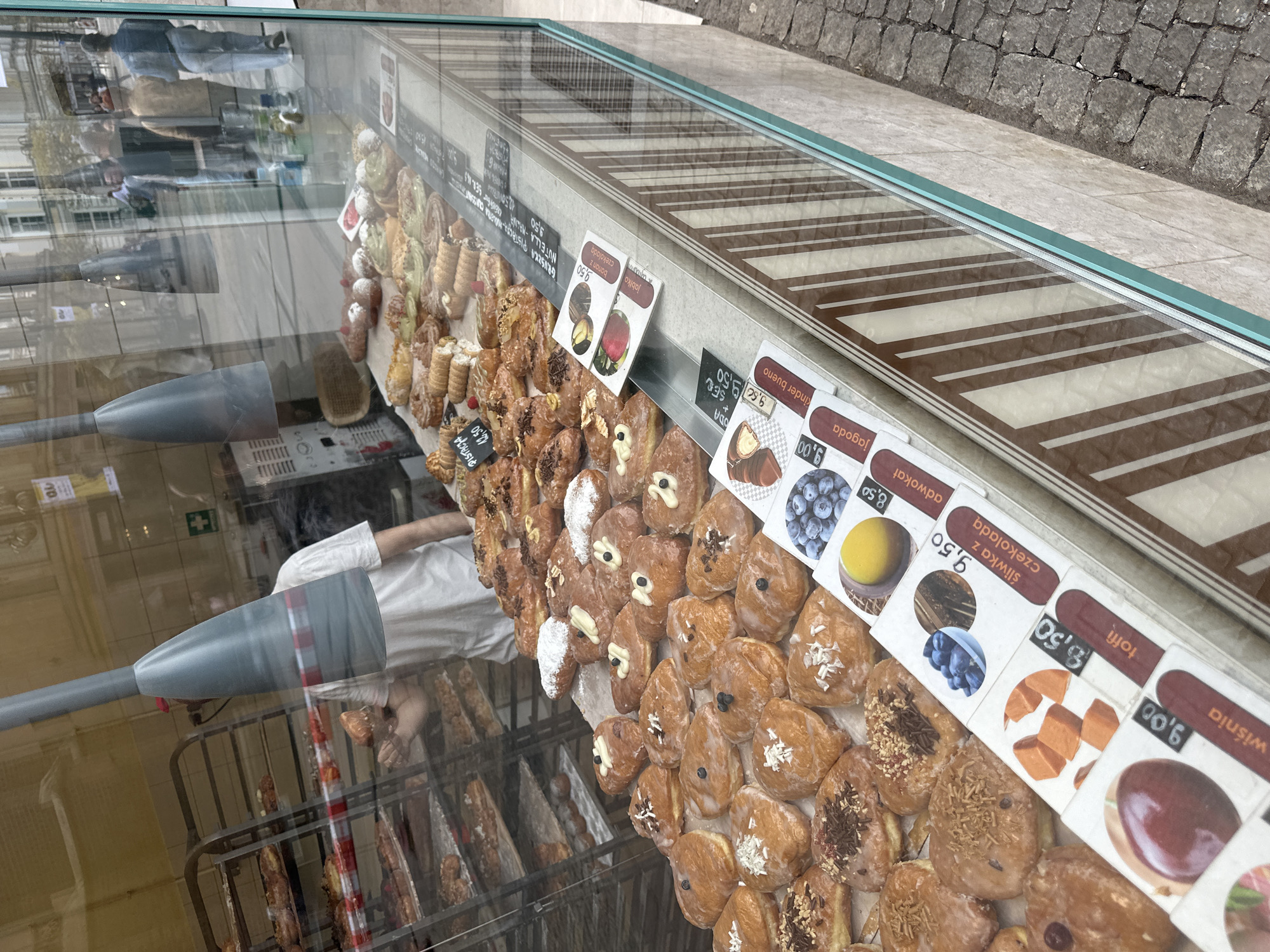 Here is a description of the image:
The image shows a display case filled with a variety of Polish pastries, mostly doughnuts (pączki) and cream-filled pastries. Each type of pastry has a corresponding label with its name in Polish and its price. Based on the names on the labels, the bakery is likely located in Poland, specifically in the city of Wrocław, as “Wiśnia” is Polish for cherry. The top of the display case is reflective, showing a reflection of the street and passersby. Inside the bakery, a person wearing a white shirt is visible. Behind the pastry display is a metal rack holding more pastries. The overall scene suggests a traditional Polish bakery.