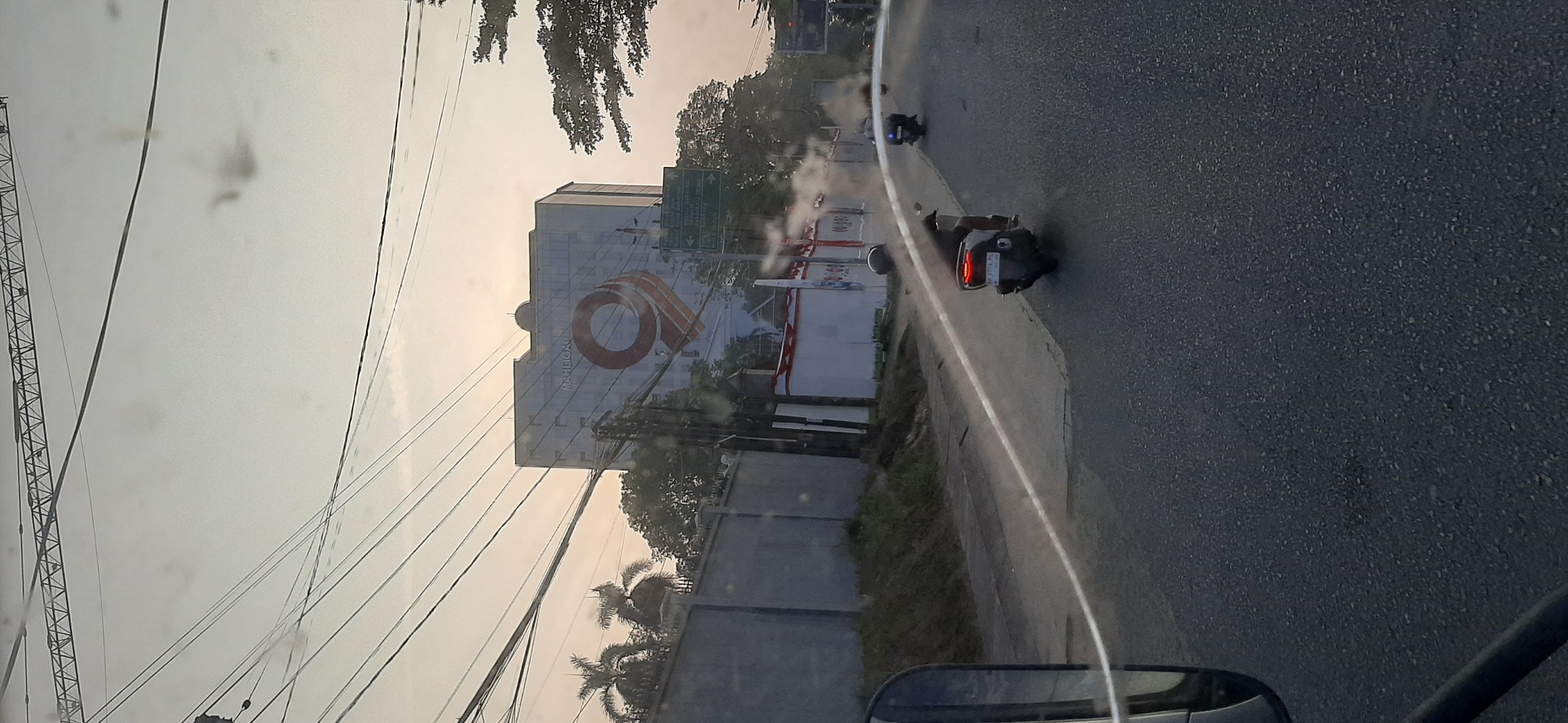 The image depicts a street scene in Jambi, Indonesia. In the background, a modern, light-colored building is prominently visible, featuring a large logo with the number "9" and the word "MAHLIGAI" in text. The building is situated behind a grey concrete wall topped with a fence, and palm trees are visible to the left of the building. A network of power lines crisscrosses the sky above, with a large metal structure, possibly part of a crane or overhead infrastructure, also present.
The street in the foreground is paved with asphalt. Several motorcycles are in motion on the road. One dark-colored scooter is positioned in the right lane, with its brake light illuminated. Another motorcycle, slightly further ahead, is also visible. The perspective suggests the photo was taken from inside a vehicle, with a portion of its side mirror visible on the left edge of the frame. There are some streaks or smudges on what appears to be a windshield or camera lens. The overall lighting suggests it is daytime, possibly late afternoon, with a soft, hazy sky. Red and white decorative elements are visible on a wall to the right of the building. A green directional signpost is also present near the building.