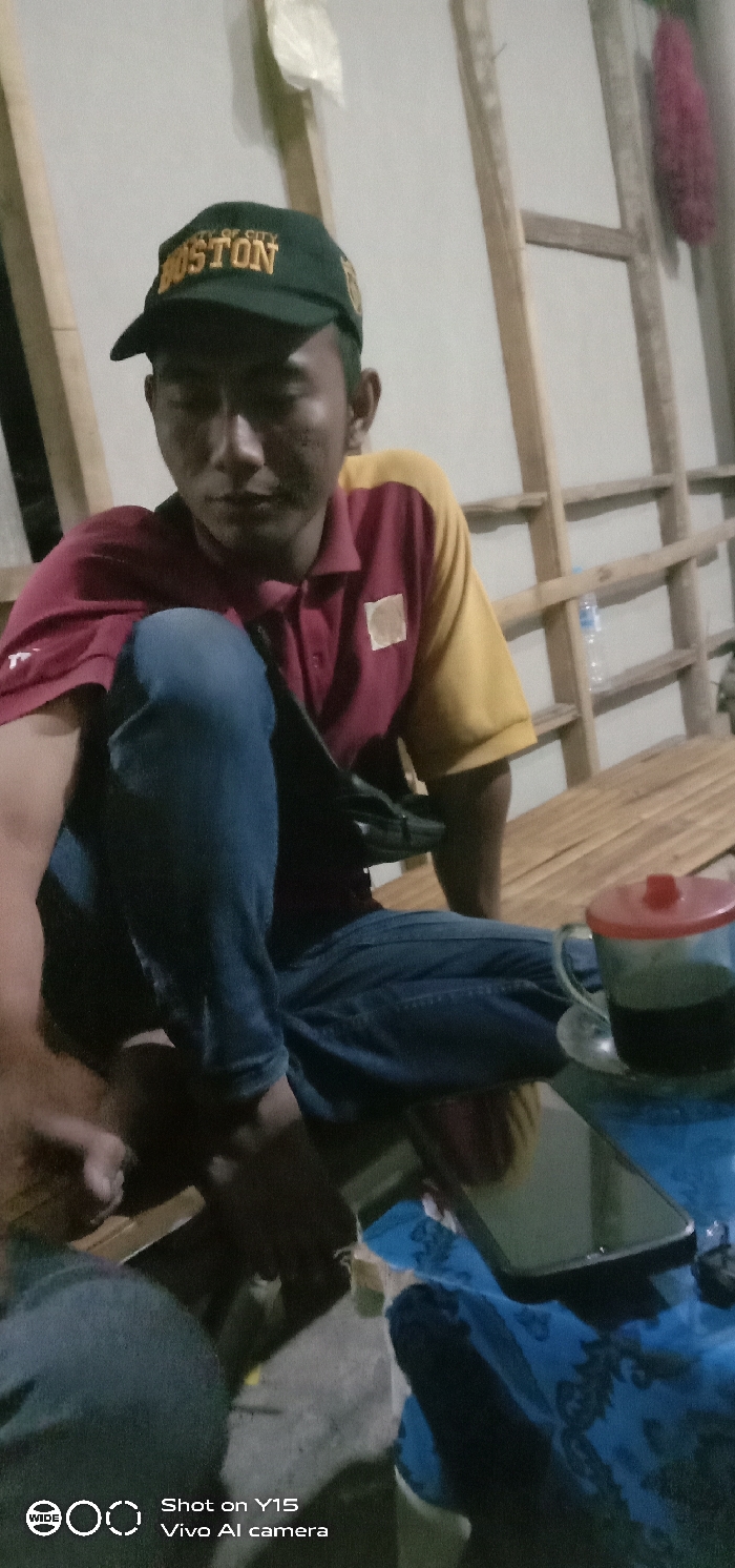 This image depicts a male individual seated indoors in a location within the city of Jabungwetan, Indonesia. The man wears a dark green baseball cap with "BOSTON" embroidered in yellow, a two-toned polo shirt featuring a maroon body and yellow sleeves, and blue jeans. A black sling bag is visible across his torso. His head is tilted downwards and slightly to the left. To his right, on a blue surface, a clear glass mug containing a dark liquid and fitted with a red plastic lid rests next to a black smartphone. The background features a light-colored wooden slat wall construction against a pale wall. A clear plastic water bottle, a white plastic bag, and a red fibrous object are also visible hanging on the background structure. The individual is positioned on a surface composed of light-colored parallel slats.