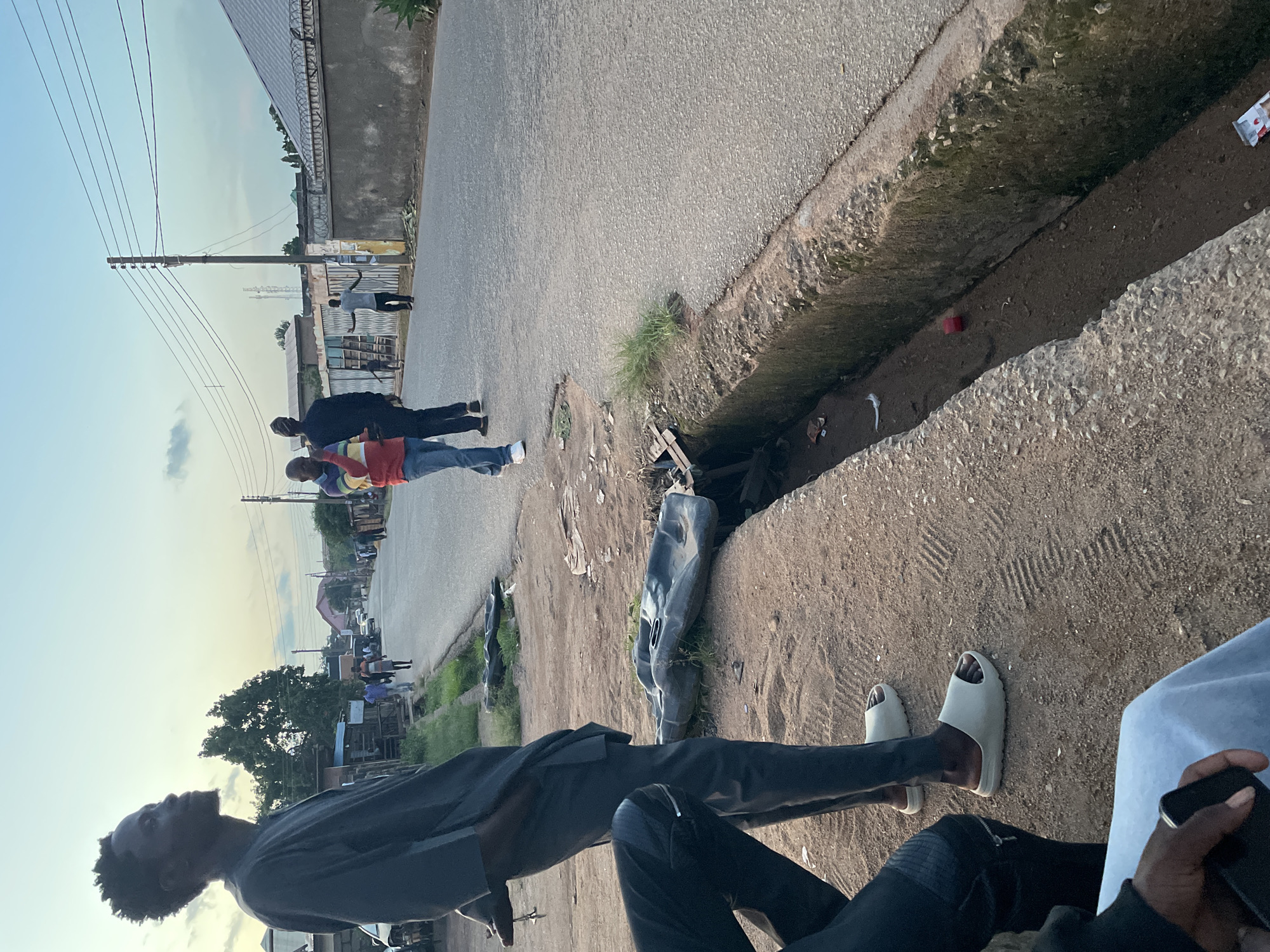 The image depicts an outdoor street scene in Jos, Nigeria, under a bright, light blue sky with scattered clouds.
In the foreground, positioned to the left, a dark-skinned male with short, dark, textured hair stands in profile, gazing rightward into the distance. He wears a dark grey, short-sleeved kaftan-style tunic over matching trousers and light grey slide sandals. His right hand holds a small metallic object, possibly keys. Partially visible at the bottom left is the left leg of another individual, clad in dark trousers with a zippered pocket, and a dark-skinned hand holding a black mobile phone over light blue fabric.
The scene features an asphalt road running centrally, flanked by a dirt and sparse-grass shoulder on the right. An open, concrete drainage ditch, partially filled with earth and minor refuse including a red cap and a white wrapper, runs parallel to the road on the right. Two black, irregularly shaped plastic objects, resembling automotive components, rest on the dirt shoulder next to the ditch.
On the road in the mid-ground, a male in a multi-colored striped polo shirt (red, yellow, blue, white) and blue jeans walks left, partially obscured by another male in dark attire walking away from the camera. Further down the road, another male in a light shirt and dark trousers stands with both arms extended horizontally.
A wooden utility pole with multiple overhead electrical or communication wires stands on the right side of the road. Beyond the pole, a grey concrete wall topped with barbed wire fencing stretches into the distance. The background features various low-rise buildings with corrugated roofs and additional fencing, along with a prominent tree on the left side of the road. Several other indistinct figures are visible walking in the distance along the road.