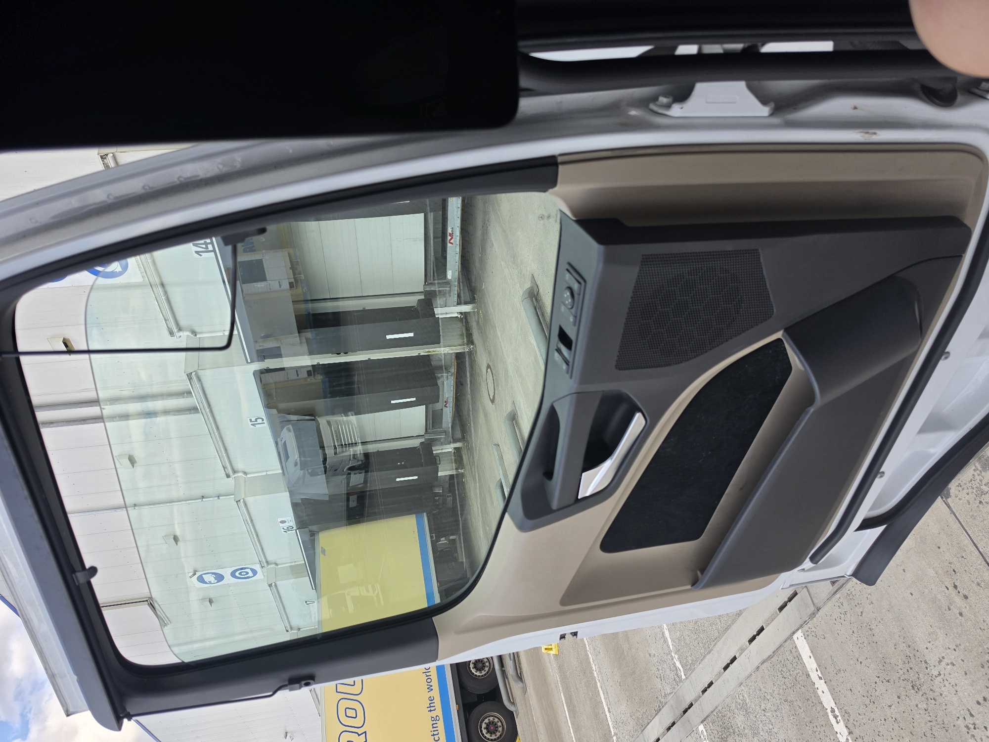 The image captures the interior of a vehicle, likely a truck or van, with the driver's side door open. The perspective is from inside the cabin, looking out through the open door and the side window.
The interior of the door is visible, featuring a dark grey door panel with a speaker grille, a chrome door handle, and a textured fabric insert. The upper part of the doorframe and window seal are also visible, along with the outer body of the vehicle.
Through the open door and the glass, a reflection of the exterior is seen. This reflection shows a row of large white industrial doors, possibly for a warehouse or loading bay, with numbers "14", "15", "16", and "17" painted above them. There are also some signs with symbols, including one depicting a person with a backpack and another with a car and a prohibited sign. In the foreground of the reflection, a yellow truck with blue lettering is partially visible, with its wheels and part of the trailer showing.
The ground outside appears to be a concrete surface, with some white markings. The lighting suggests it is daytime, and the sky is visible in the upper left corner, showing some clouds.
The scene seems to be an industrial or logistics area, given the presence of the warehouse-like structures and the truck. There is no indication of people or any specific activity taking place beyond what is implied by the setting. The overall impression is that of a paused moment, perhaps before or after loading/unloading operations.
The visible text includes numbers on the building ("14", "15", "16", "17") and lettering on the yellow truck, which appears to include "ROL" and "ecting the worl".