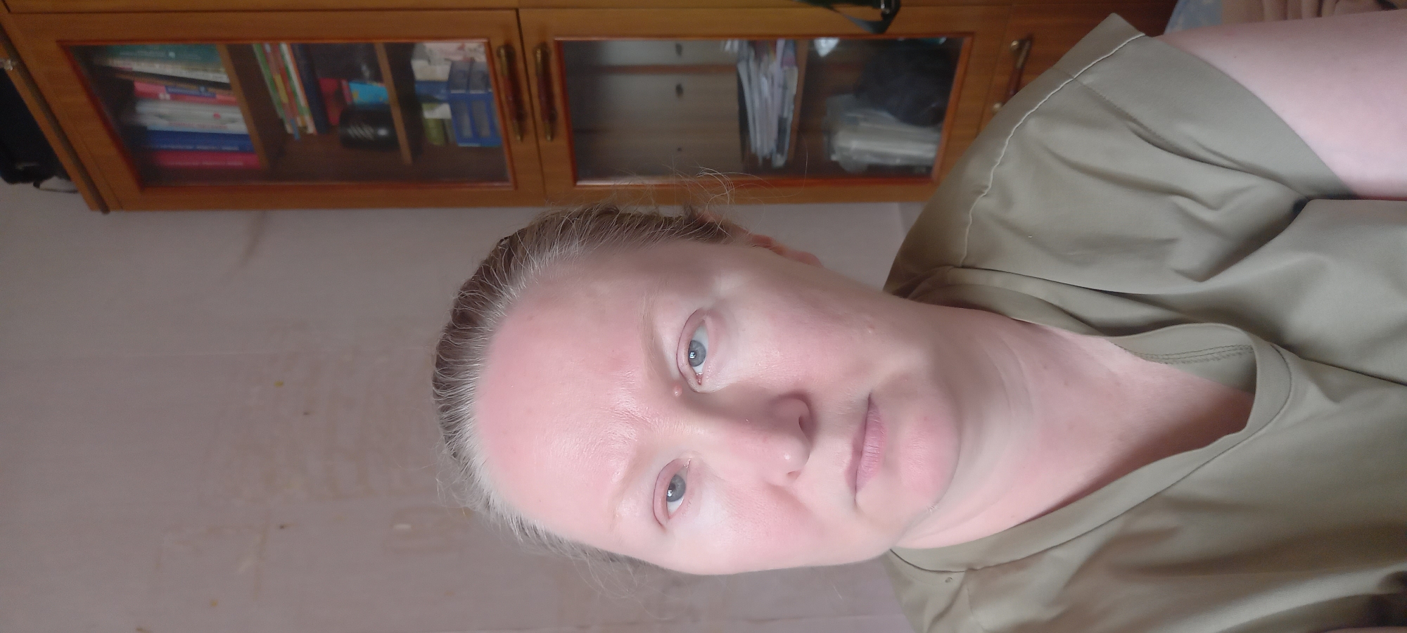 The media file shows a close-up, upward-facing selfie of a fair-skinned woman with light blonde hair pulled back from her face. She has blue-grey eyes, a light dusting of freckles, and appears to be in a casual setting. She is wearing a khaki-colored shirt, and her skin appears slightly flushed. The background is out of focus but suggests an indoor environment with a wooden cabinet or bookshelf containing items, possibly books or files. The lighting appears to be natural, suggesting it could be daytime. The woman's expression is neutral, with a slight tilt of her head. The overall mood is casual and informal. The image is oriented sideways. There is no visible text in the image. The location context provided is Izhevsk, Russia.