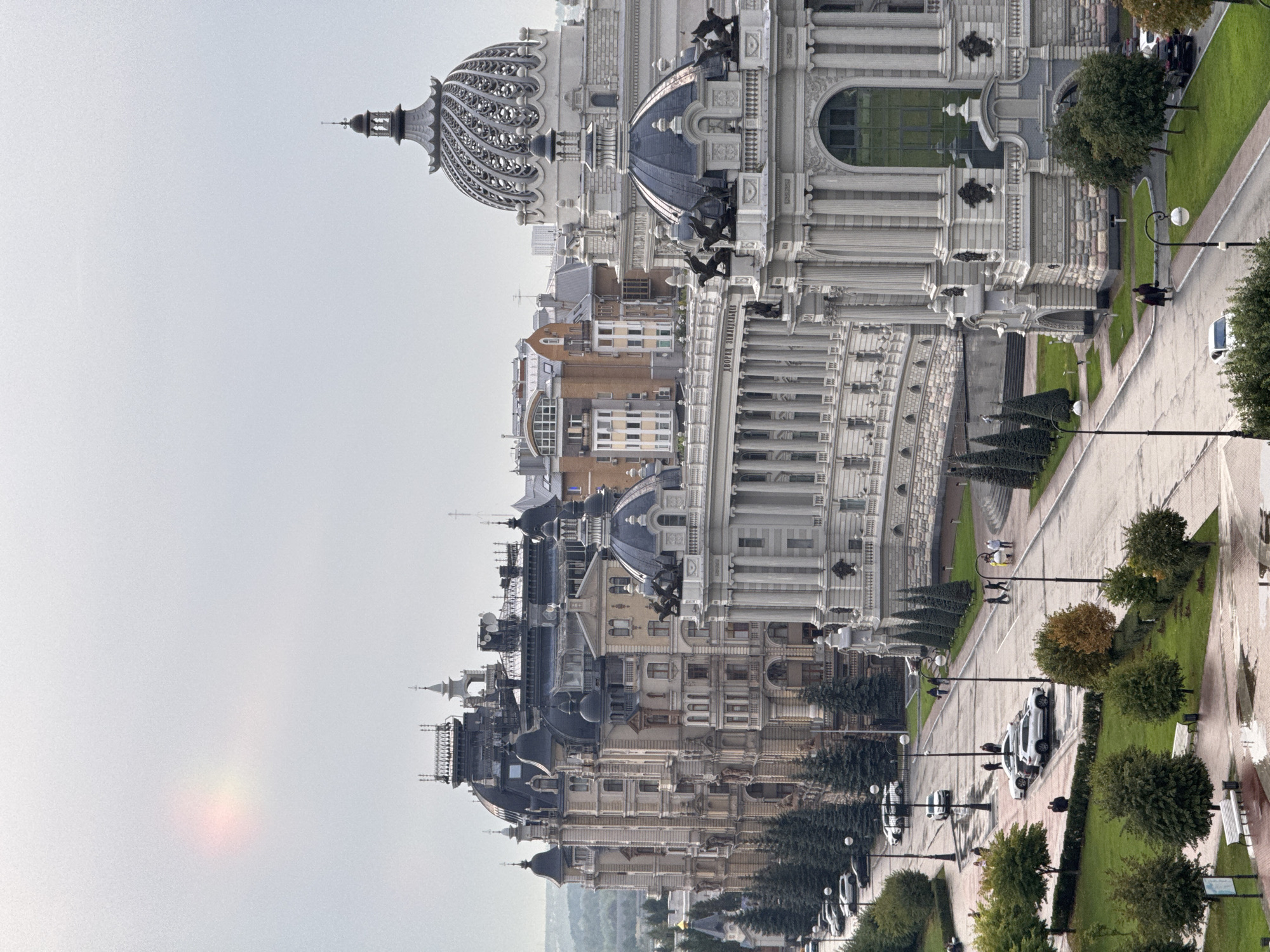 The image depicts a street scene in Kazan, Russia, featuring the Palace of Agriculture and the National Library of the Republic of Tatarstan. The architecture is grand and ornate, with classical elements like columns and domes. Several buildings line the street, showcasing intricate stonework and multiple levels. Cars are parked along the street and some are in motion. A few people are walking along the street and on the sidewalks. The sky is overcast, with a faint, colorful light anomaly in the upper left corner, possibly a lens flare or atmospheric phenomenon. Trees and green spaces are visible along the street, and the ground appears wet, suggesting recent rain.