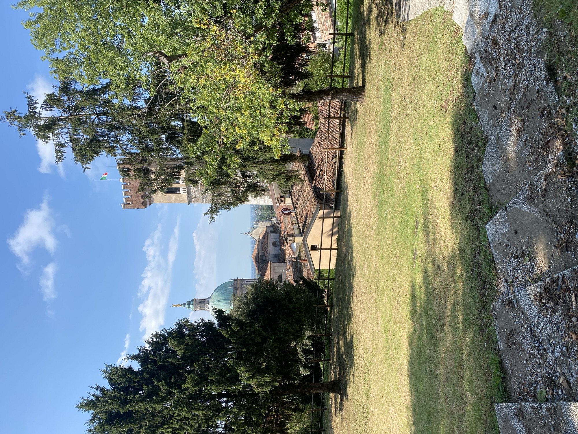 The image shows a scenic view of Lonato del Garda, Italy, dominated by the imposing Rocca Viscontea, a medieval fortress with a prominent tower flying the Italian flag. In the foreground, a well-maintained lawn slopes down towards a wooden fence, separating it from a cluster of terracotta-roofed buildings. A large, dark green evergreen tree stands to the left, while a smaller deciduous tree with yellowing leaves is positioned to the right, casting shadows on the grass. In the background, a majestic green dome, likely part of a church or cathedral, rises above the rooftops, topped with a golden statue. The sky is a clear blue with scattered white clouds. Stone steps, partially covered in gravel and fallen leaves, lead down into the scene from the bottom right corner. The overall impression is one of a serene, sun-drenched afternoon in a historic Italian town.