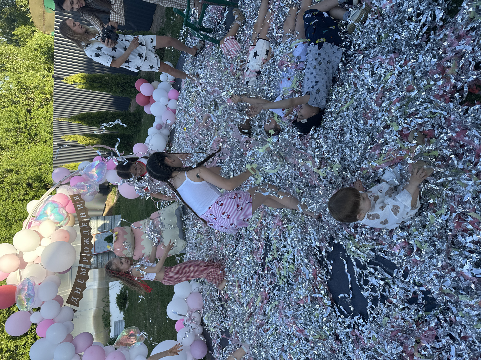 The image captures a birthday celebration, likely taking place outdoors, possibly in a backyard in Kazan, Russia, indicated by the Cyrillic inscription "Днём рождения" (Happy Birthday) on a sign adorned with pink and white balloons. Several children are lying on a ground covered in silver and pink confetti, seemingly enjoying themselves amidst the celebratory mess. Their attire includes casual wear such as pajamas, shorts, and tank tops. An adult woman, wearing star-patterned pajamas and holding a camera, is likely documenting the event. A second adult can be seen with a checkered pattern shirt. There's a balloon arch and a birthday "cake" made of balloons. The background includes greenery such as trees and shrubs, and a fence.