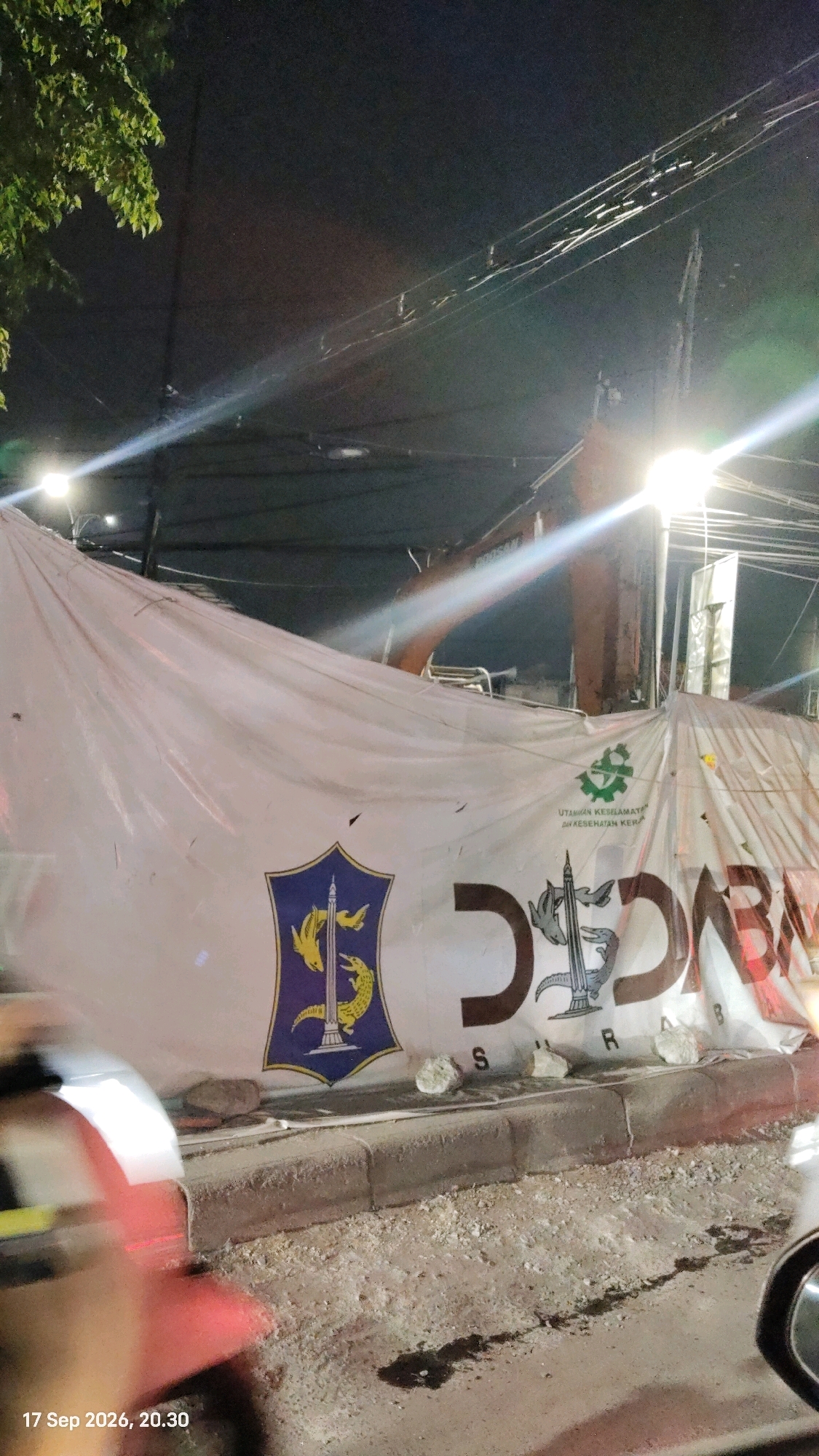 The image depicts a construction site at night, with a large white tarp covering a section of the area. The tarp features several emblems and text. On the left, there is a blue and yellow shield with an image of a building and two figures. To the right of this shield, there is a green circular emblem with the text "UTAMAKAN KESELAMATAN DAN KESEHATAN KERJA" above it. Further to the right, large dark letters appear to spell out "DDABAR," though they are partially obscured and stylized. Scattered on the ground in front of the tarp are several rocks and debris. A construction vehicle, likely an excavator, is visible behind the tarp. Overhead, numerous electrical wires crisscross the scene, illuminated by bright streetlights, creating lens flares. The foreground shows a curb and the ground covered in dirt and some dark liquid spills. A blurred red object, possibly part of a vehicle, is visible on the far left. The timestamp "17 Sep 2026, 20.30" is present in the bottom left corner. The scene suggests a road construction or renovation project in Surabaya, Indonesia.