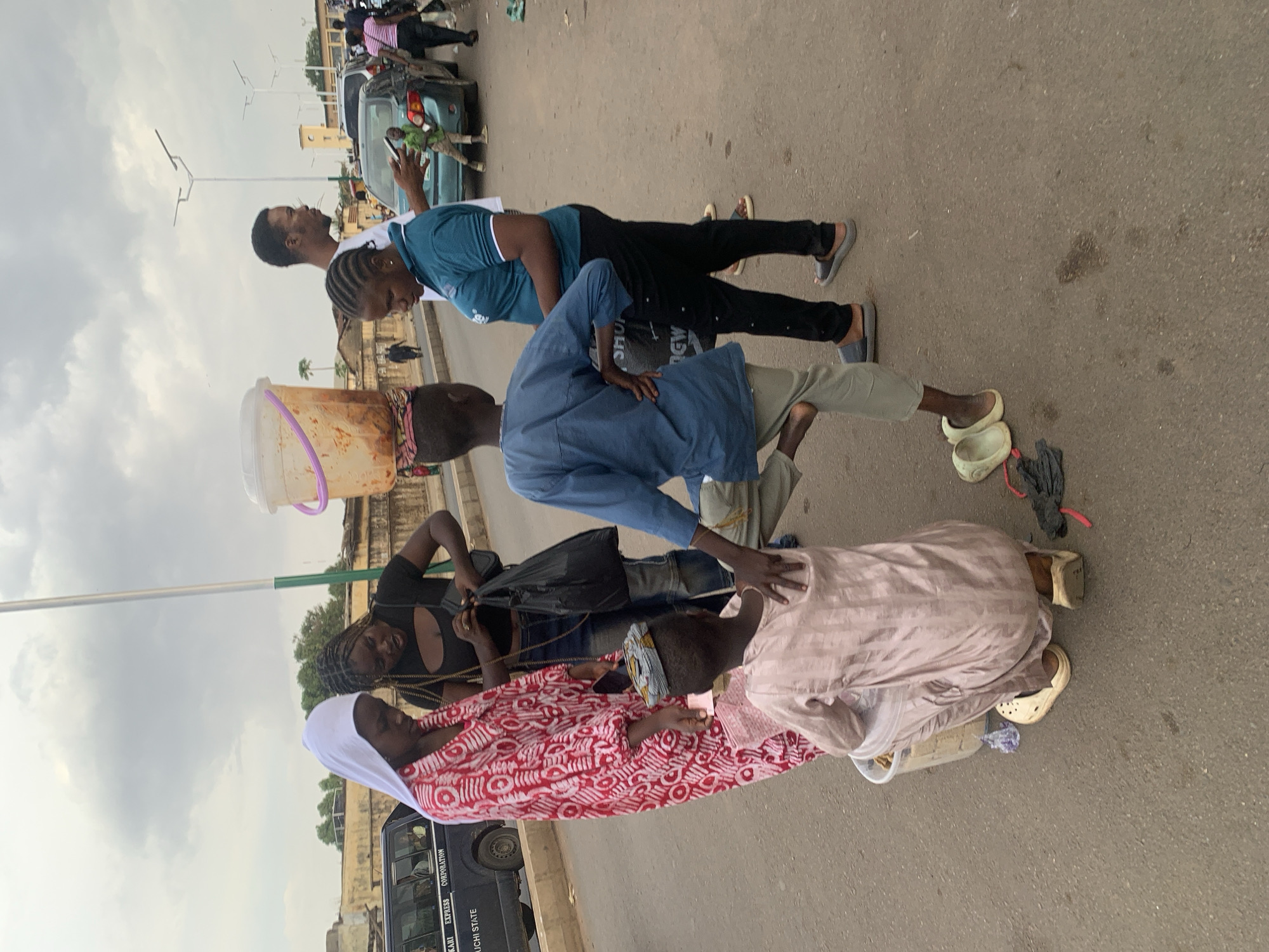 The image depicts an active street scene in Jos, Nigeria, under an overcast sky during daytime. A gathering of people is concentrated on a paved road, where a man in a blue t-shirt and black trousers is performing what appears to be a plank or push-up. Several individuals are observing this activity intently. A man in a light blue shirt leans forward, looking down at the man on the ground. Beside him, a woman in a red and white patterned dress and a white headscarf is seated or kneeling, holding a mobile phone, possibly recording the event. Another person in a black top also stands nearby, gazing downwards.
Further back, a person balances a large, clear plastic bucket, which seems to contain an orange-red substance, on their head, either observing the scene or passing through. The background features more pedestrians, parked vehicles including a light blue car, and a dark-colored bus. Visible text on the side of the bus includes "KAI EXPRESS CORPORATION" and "NAUCHI STATE". The overall impression is that of a public street performance or demonstration capturing the attention and curiosity of a crowd.