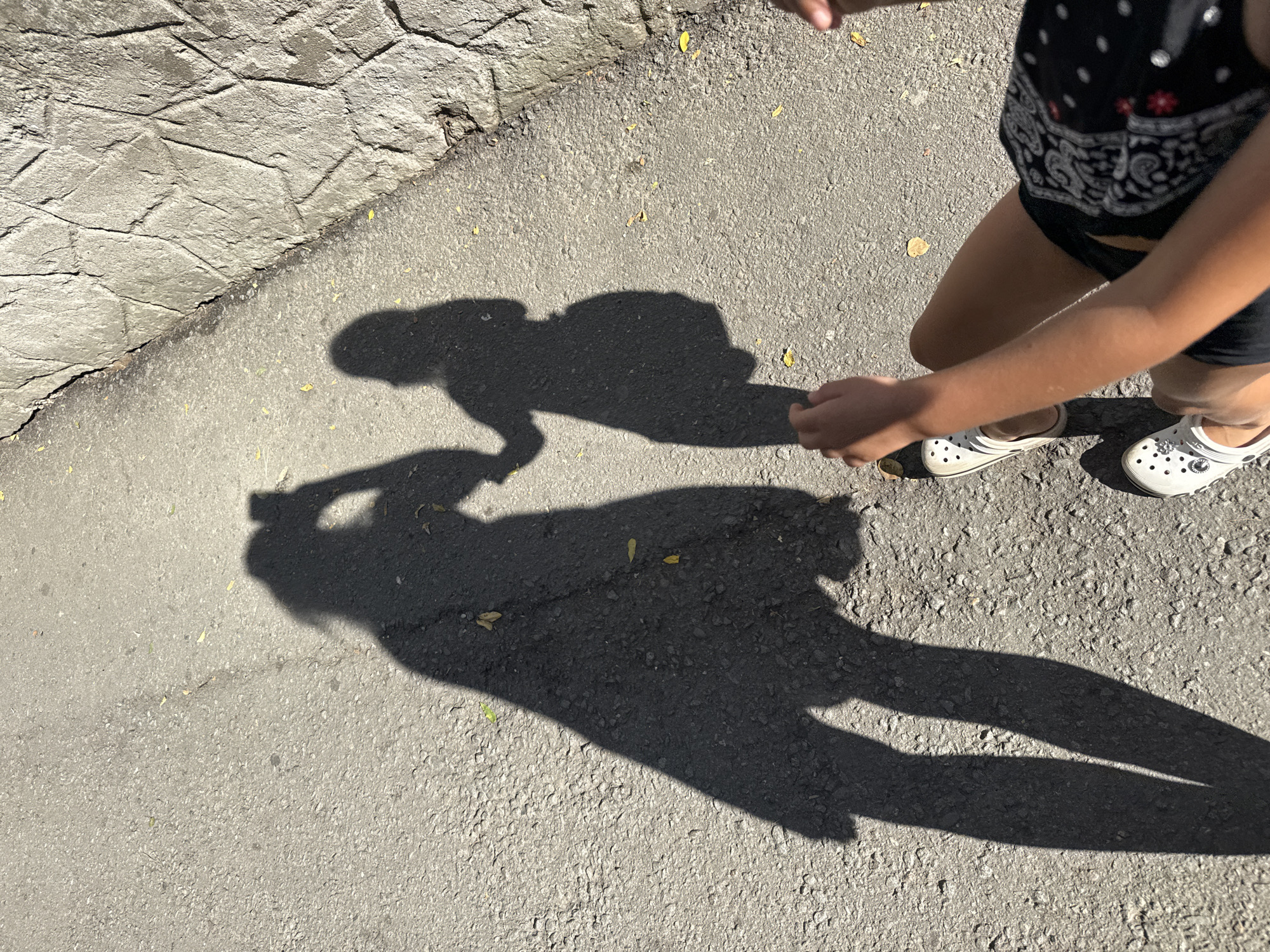 In Partenit, Ukraine, two shadows are cast on an asphalt surface adjacent to a stone-lined embankment. The shadow on the left depicts a figure holding a phone, presumably taking the photograph, with their arm extended. To the right of this figure, a second shadow, appearing smaller and potentially representing a child, is holding hands with the first figure. The perspective suggests the photograph is taken from a high angle, looking down at the shadows and the ground. The ground is a mix of asphalt and a rough, textured stone wall. Scattered yellow leaves are visible on the asphalt. In the foreground, the lower legs and feet of a person wearing white clogs are visible, along with a portion of their torso clad in a black garment with a white pattern. The shadows indicate a bright, sunny day.