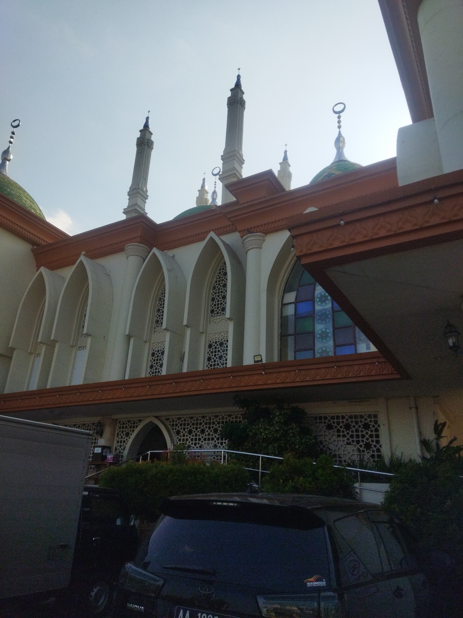 The image captures a low-angle view of the exterior of the Grand Mosque of Sukoharjo, located in Sukoharjo, Indonesia. The mosque features multiple white minarets topped with crescent moons, green domes, and decorative arched windows with stained glass. The building's facade is adorned with ornate detailing in brown and white. In the foreground, a dark-colored SUV and a gray box truck are parked, partially obscuring the lower portion of the mosque. Lush green foliage surrounds the base of the building. The sky is a pale, overcast blue.