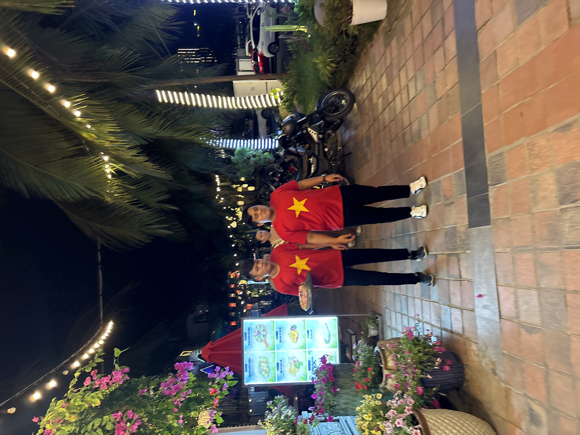 This nighttime image, captured in Đà Nẵng, Vietnam, features two young males standing on a brick pavement. Both are wearing identical red t-shirts adorned with a prominent yellow star, reminiscent of the Vietnamese flag, paired with black pants and white athletic shoes. The person in the foreground appears to be holding a plate or tray.
The setting is an outdoor area, possibly a promenade or market street, characterized by abundant tropical flora including large palm trees and potted plants with vibrant pink, purple, and yellow flowers. The scene is warmly lit by numerous string lights draped across the palm fronds and structures. In the background, parked motorcycles and a white car are visible, along with other indistinct figures.
A brightly illuminated menu board stands prominently, displaying images of seafood dishes. The visible Vietnamese text from the board includes: "TÔM HÙM NƯỚNG PHOMAI" (Grilled Lobster with Cheese), "TÚ HÀI NƯƠNG MỠ HÀNH" (Grilled Sea Anemone with Scallion Oil), "TÔM VẸ CHẤY TỎI" (Sautéed Shrimp with Garlic), and "TÔM HÒM SỐT ĐỘ TỎI" (Lobster with Garlic Sauce). The pavement consists of red-brown bricks interspersed with a dark grey central stripe. The overall atmosphere suggests a casual, lively evening at a food establishment in a tropical climate.