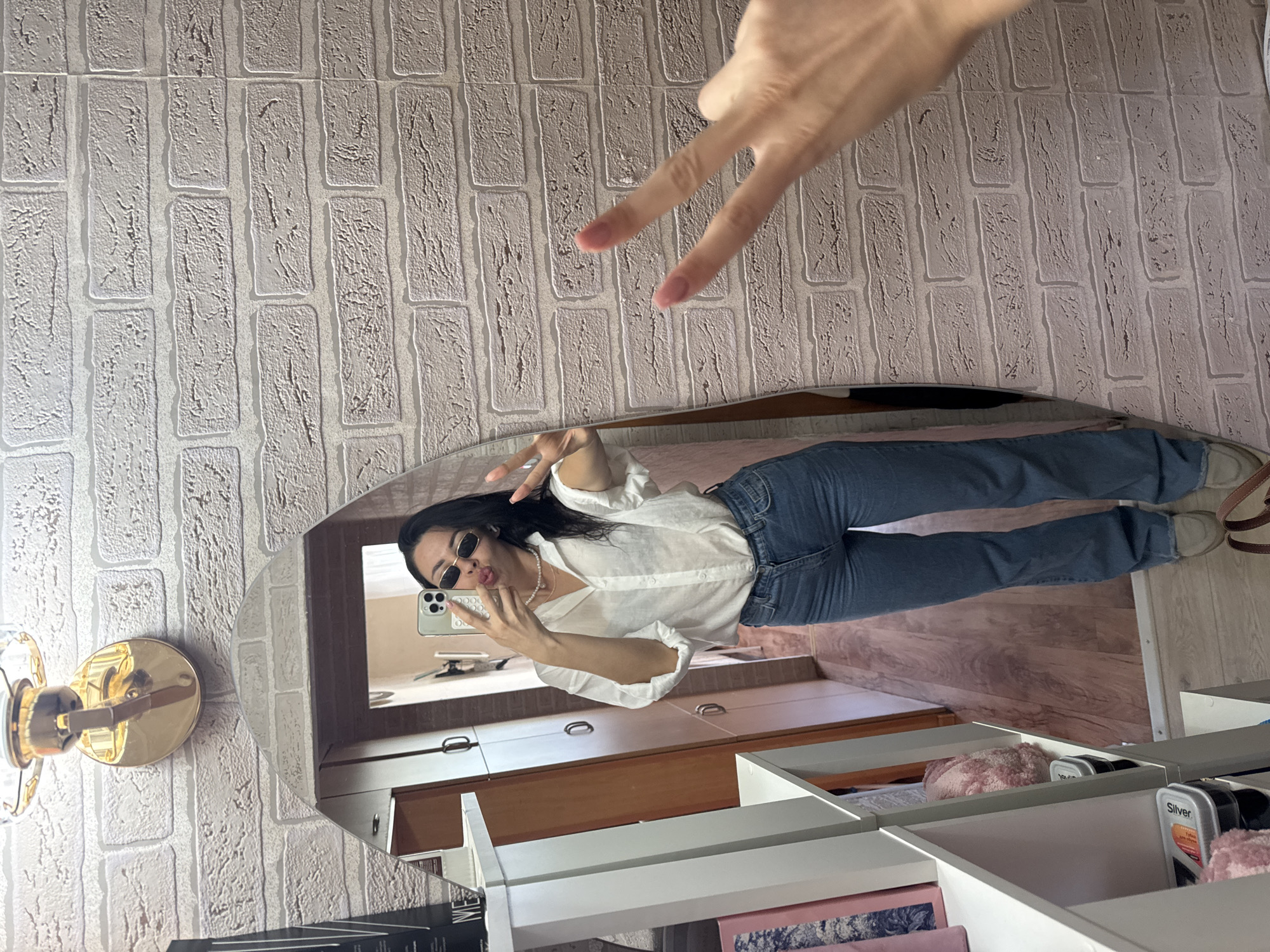 A woman stands in front of an oval mirror, taking a selfie. She is wearing a white button-down shirt, blue jeans, and sunglasses. She has her hair down and is wearing a pearl necklace. She is holding up two fingers in a peace sign with one hand and her phone in the other. The background is a brick-patterned wallpaper. There is a shelf to the left of the mirror with some items on it. The scene is indoors, likely in a residential setting. There is no indication of the city of Chaikovsky, Russia in the image.