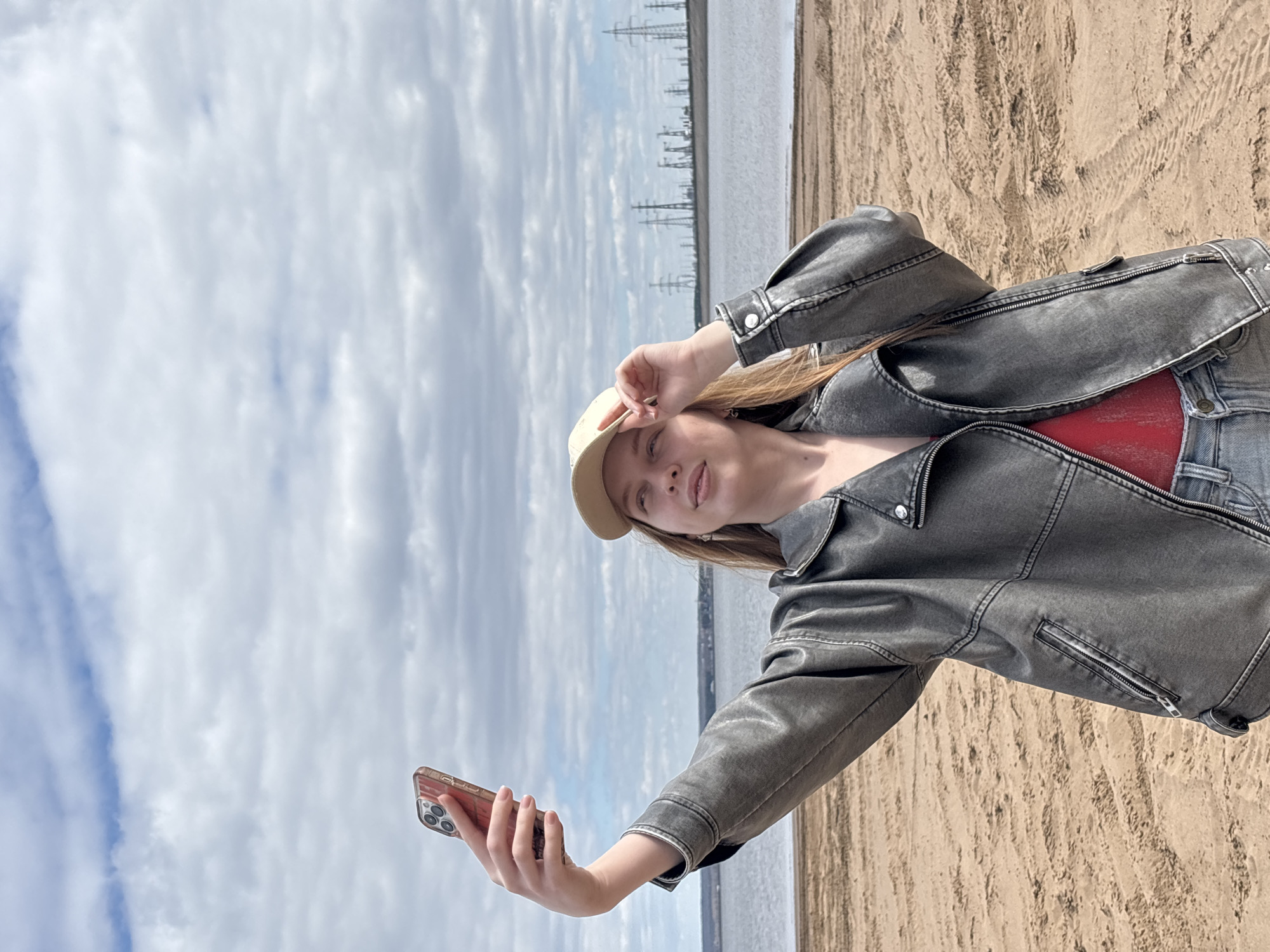 The image depicts a woman taking a selfie on a beach. She is wearing a beige baseball cap, a gray leather jacket over a red shirt, and blue jeans. She is holding a smartphone in her right hand, with her left hand near her forehead. In the background, there is a body of water and power lines stretching across the horizon. The beach is sandy, with tire tracks visible in the sand. The sky is cloudy. The location is likely in Noviy, Russia, based on the metadata.