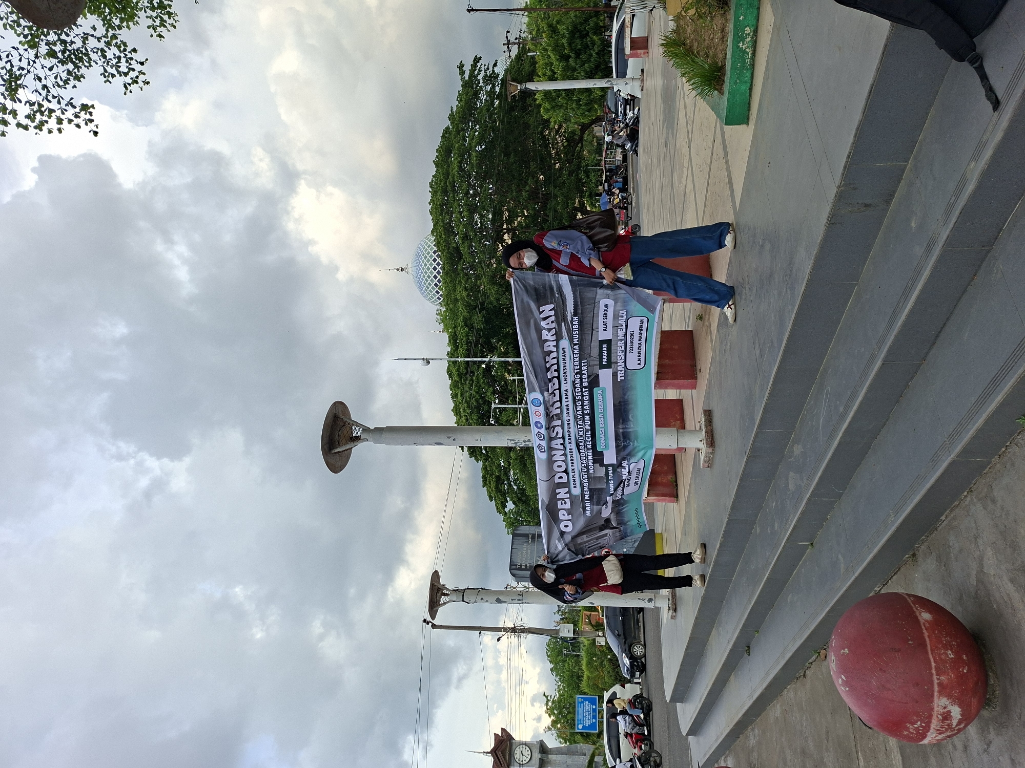 Two individuals stand on stairs, holding a banner that reads "OPEN DONASI KEBAKARAN" (Open Fire Donation). The banner also mentions the location, "KOMPLEK PARDEDE KAMPUNG JAWA LAMA-LHOKSEUMAWE" (Pardede Complex, Old Java Village, Lhokseumawe), placing this scene in Lhokseumawe, Indonesia. The banner further invites donations for those affected by fire, specifying items such as clothing and school supplies. The individuals are wearing face masks, suggesting the presence of a pandemic or health concern. In the background, there are light posts, trees, and a mosque dome. The sky is overcast. A red spherical object is near the base of the stairs.