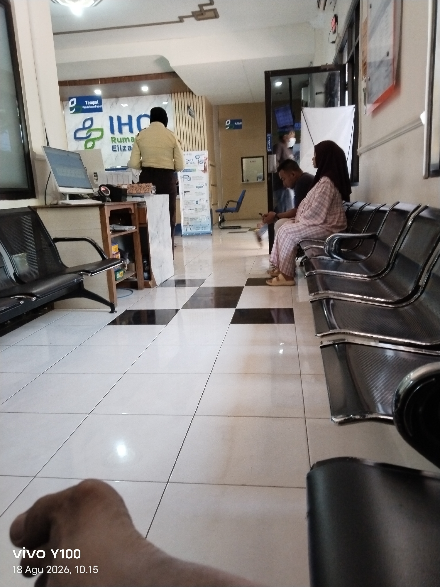 In Patokan, Indonesia, an indoor waiting area appears to be a part of a medical facility, possibly a clinic or hospital. The space features a tiled floor with a checkered pattern of black and white squares. Several rows of dark, metal-framed waiting chairs are visible, with one person, a woman wearing a patterned outfit and a headscarf, seated in one of them. Another individual, possibly male, is seated beside her. Further in the background, a person in a light-colored uniform is standing near a counter or reception desk, and a sign with the letters "IHC" and "Rumah Eliza" is prominently displayed on the wall, suggesting it is the "Eliza House" of IHC. A computer monitor and a speaker are present on a desk. A blue chair is also visible in the middle ground. The overall impression is that of a functional waiting area within a healthcare establishment.