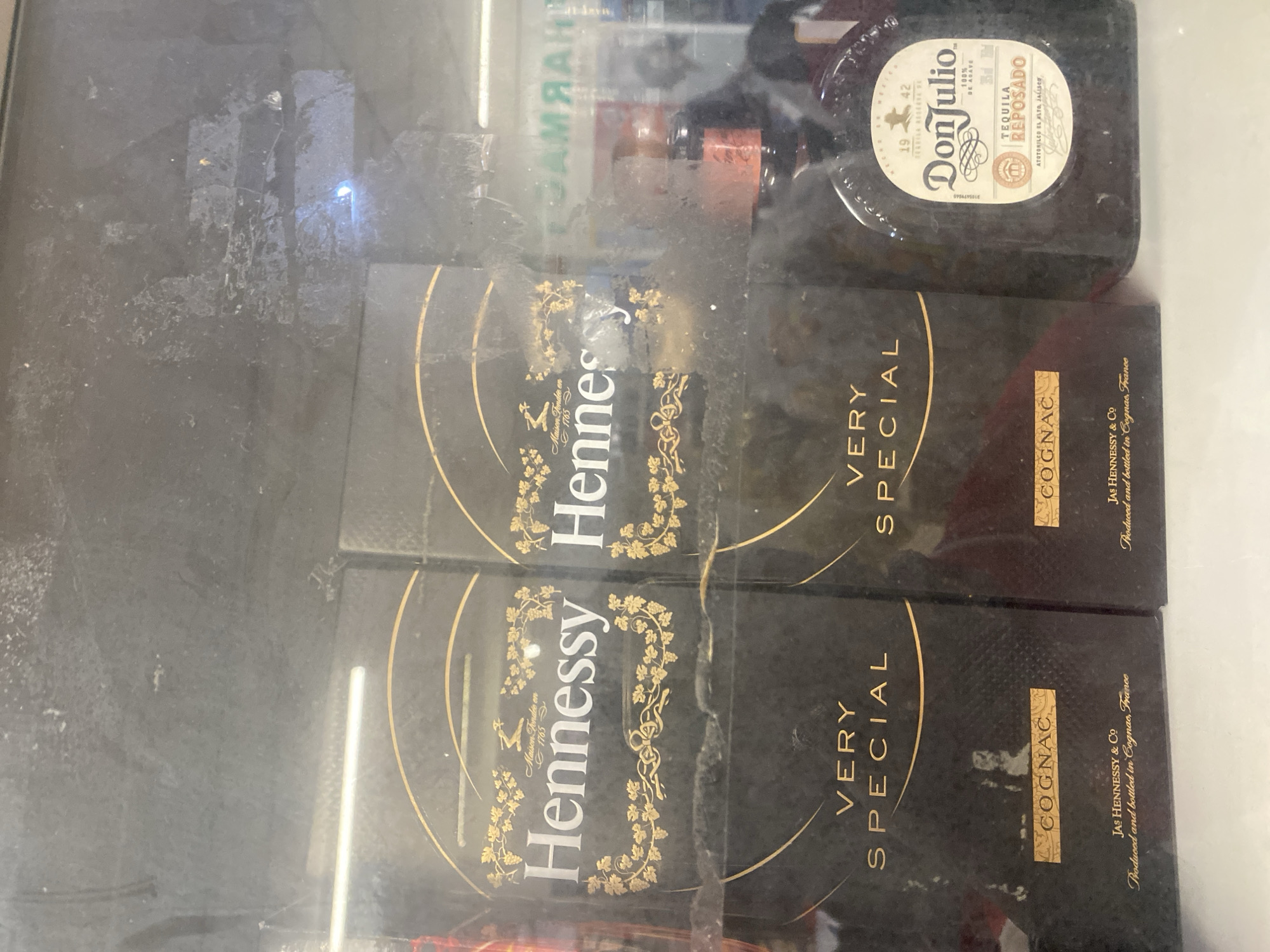 The image shows a display case with two boxes of Hennessy Very Special Cognac and a bottle of Don Julio Reposado Tequila. The boxes are dark brown with gold lettering and designs. The Hennessy boxes have "Hennessy" prominently displayed at the top, with "VERY SPECIAL" and "COGNAC" below. The bottle of Don Julio is dark glass with a cream-colored label featuring the brand name and "TEQUILA REPOSADO". The display case appears to be in a store, possibly a liquor store or supermarket, given the presence of other items and shelving in the background, including a sign that appears to read "PHARMACY". The overall scene suggests a retail environment. The image is taken from a slightly elevated angle and shows some dust or smudges on the display case glass. There are no people clearly visible in the image, but reflections suggest the presence of individuals in the vicinity. The location is identified as Ikorodu, Nigeria.