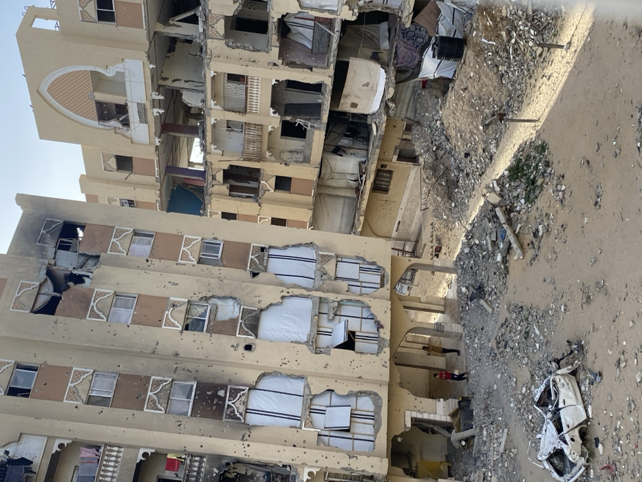 The image depicts a heavily damaged urban environment in Khan Yunis, Palestinian Territory, under a clear sky. Two multi-story buildings are prominent. The building on the left is a light-colored structure with numerous impact marks on its facade, consistent with bullet holes or shrapnel. Multiple windows are shattered or boarded with white material, and sections of the upper floors show structural collapse and charring. Below, arched open spaces lead to a ground level covered in sand and rubble. Two young males are visible standing under these arches; one wears a red shirt, the other a light brown shirt. In the foreground, a white four-door sedan is severely damaged and crushed, resting on the sandy, debris-strewn ground.
To the right and slightly behind, another multi-story building exhibits extensive structural damage and partial collapse, with multiple floors exposed and interior elements visible. Its facade is also light-colored with decorative archways. The ground between and around these buildings is covered in a dense layer of concrete rubble, sand, and various debris. Clothes are visible hanging on a balcony of the left building and draped over debris on the right building. A black water tank is partially visible amidst the debris near the base of the right building. The scene indicates significant destruction.