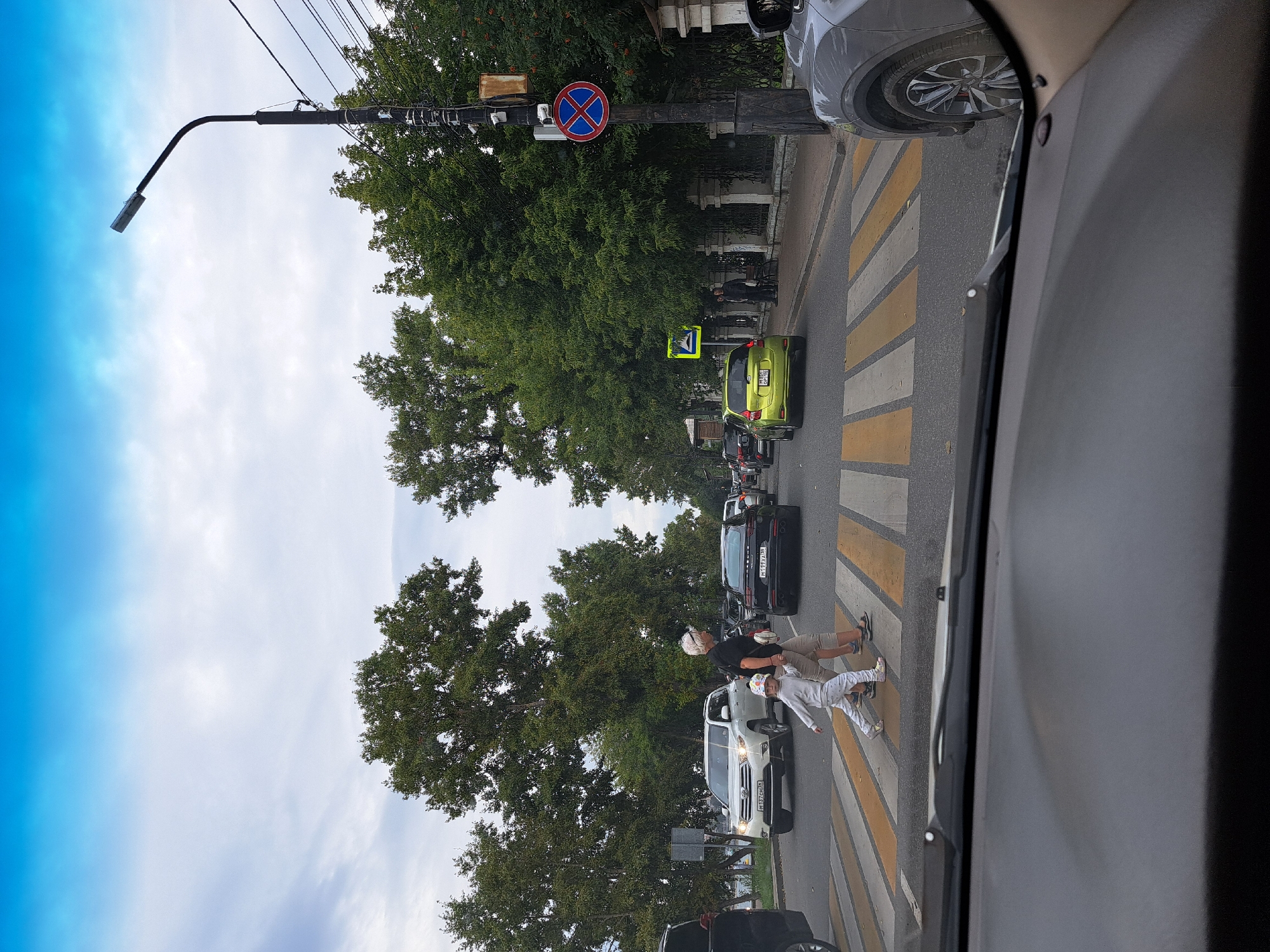 The image depicts a street scene in Irkutsk, Russia, with a pedestrian crossing in the foreground. A woman in casual attire and a child in a light-colored outfit are crossing the street from left to right. Several cars are stopped or moving in the background, including a white SUV, a black sedan, and a lime green hatchback. Trees line the street, and a lamppost with traffic signs is visible on the right side of the image. The sky is overcast with a bright blue patch at the top. The scene appears to be captured from inside another vehicle, with the dashboard visible at the bottom of the frame.