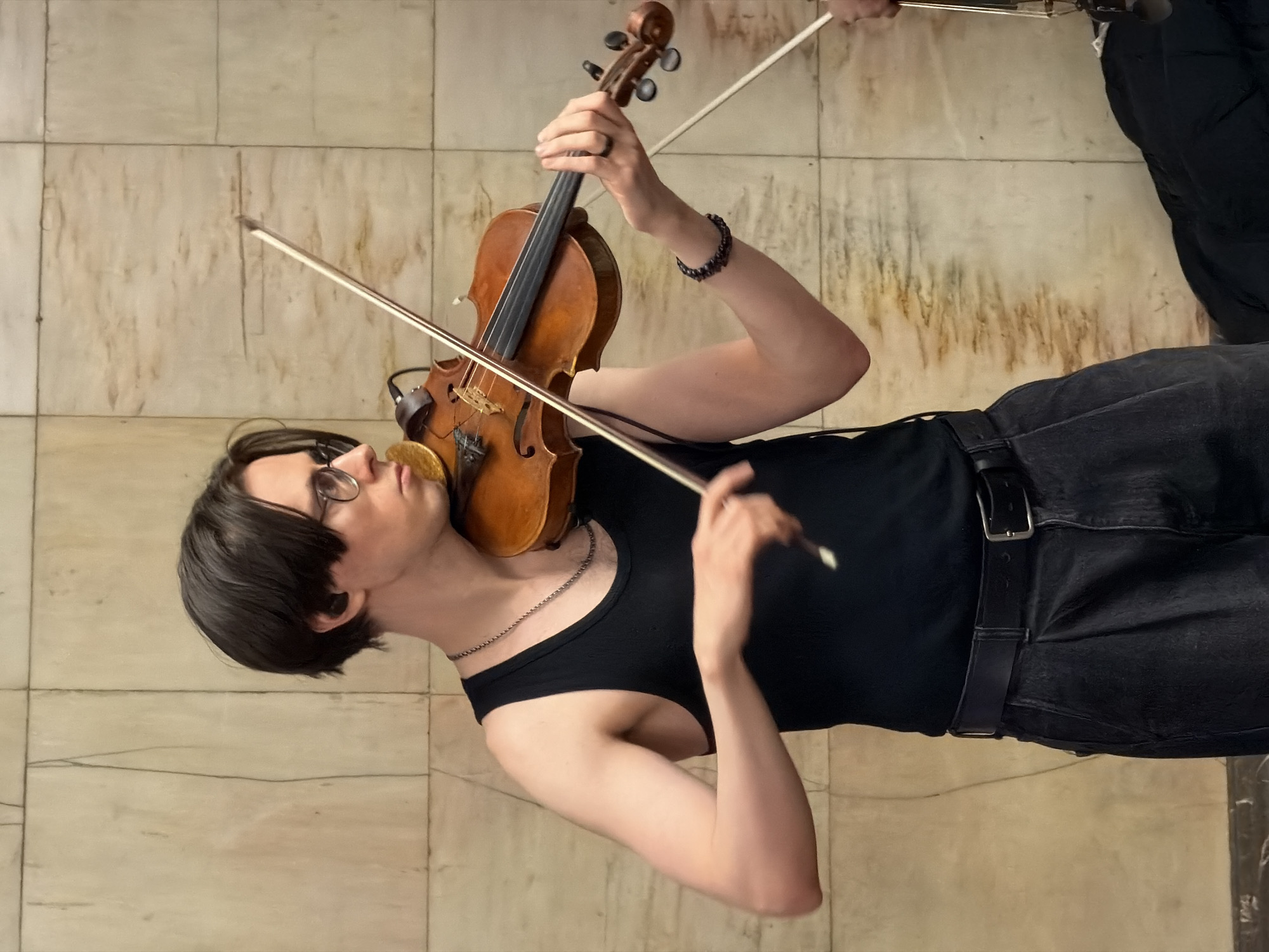 A person is playing the violin in what appears to be an indoor location. The individual, wearing glasses and a black tank top, holds the violin with their left hand and uses the bow with their right. They are also wearing black jeans and a belt. The backdrop suggests an interior with a tiled wall. The location is not identified as Moscow, Russia in the provided image.