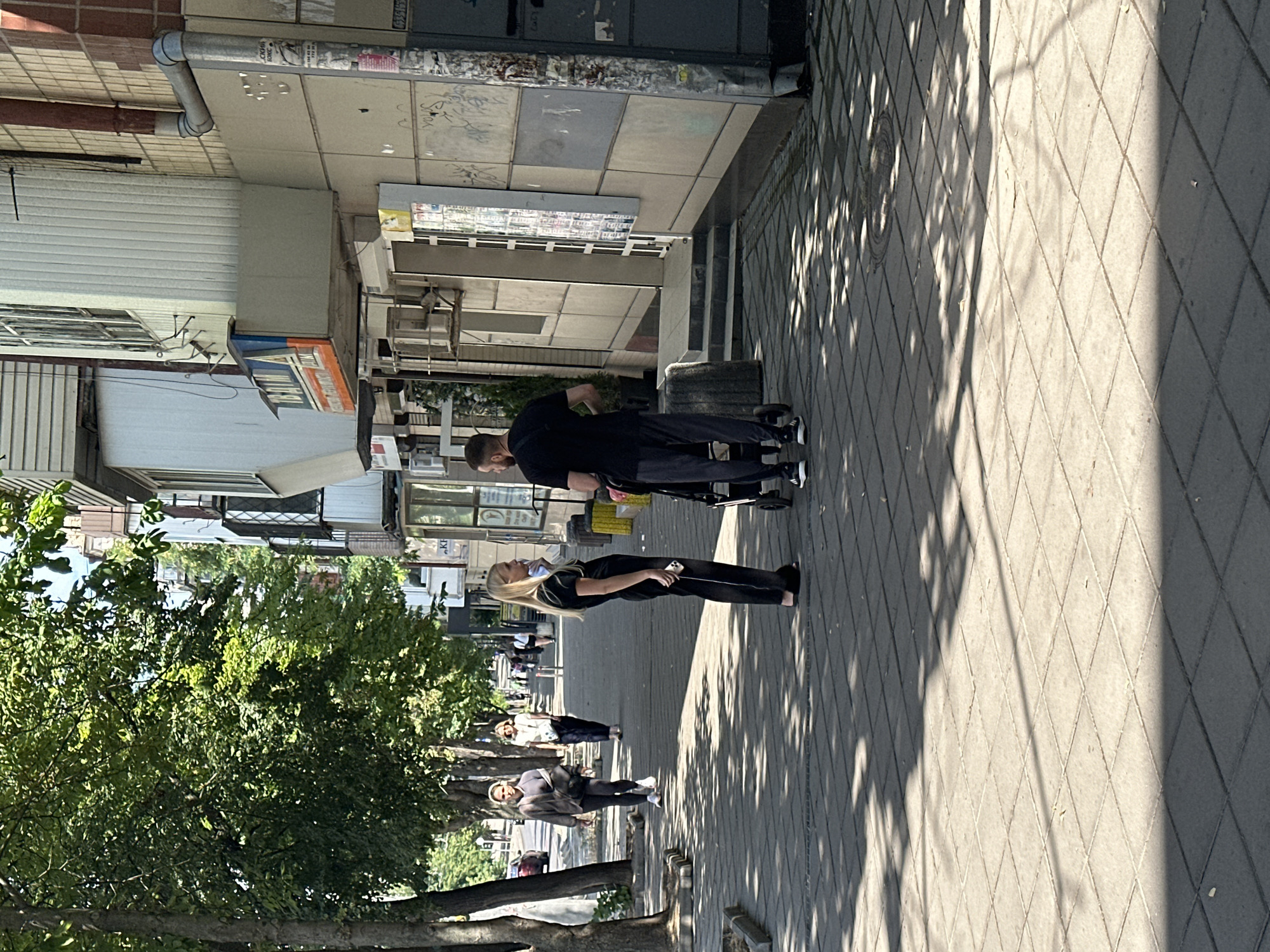 The image depicts a sun-drenched sidewalk in Odesa, Ukraine, lined with trees and a building. In the foreground, a man and a woman stand beside a stroller, engaged in conversation. The man, wearing a black t-shirt and dark pants, is looking away from the camera. The woman, dressed in black, is facing away, holding her phone. Further down the sidewalk, two other women are walking, one in a grey tracksuit and the other in a light-colored shirt and dark pants. The building features a beige facade with windows and a prominent sign. Shadows from the trees are cast across the tiled pavement, indicating it is daytime.