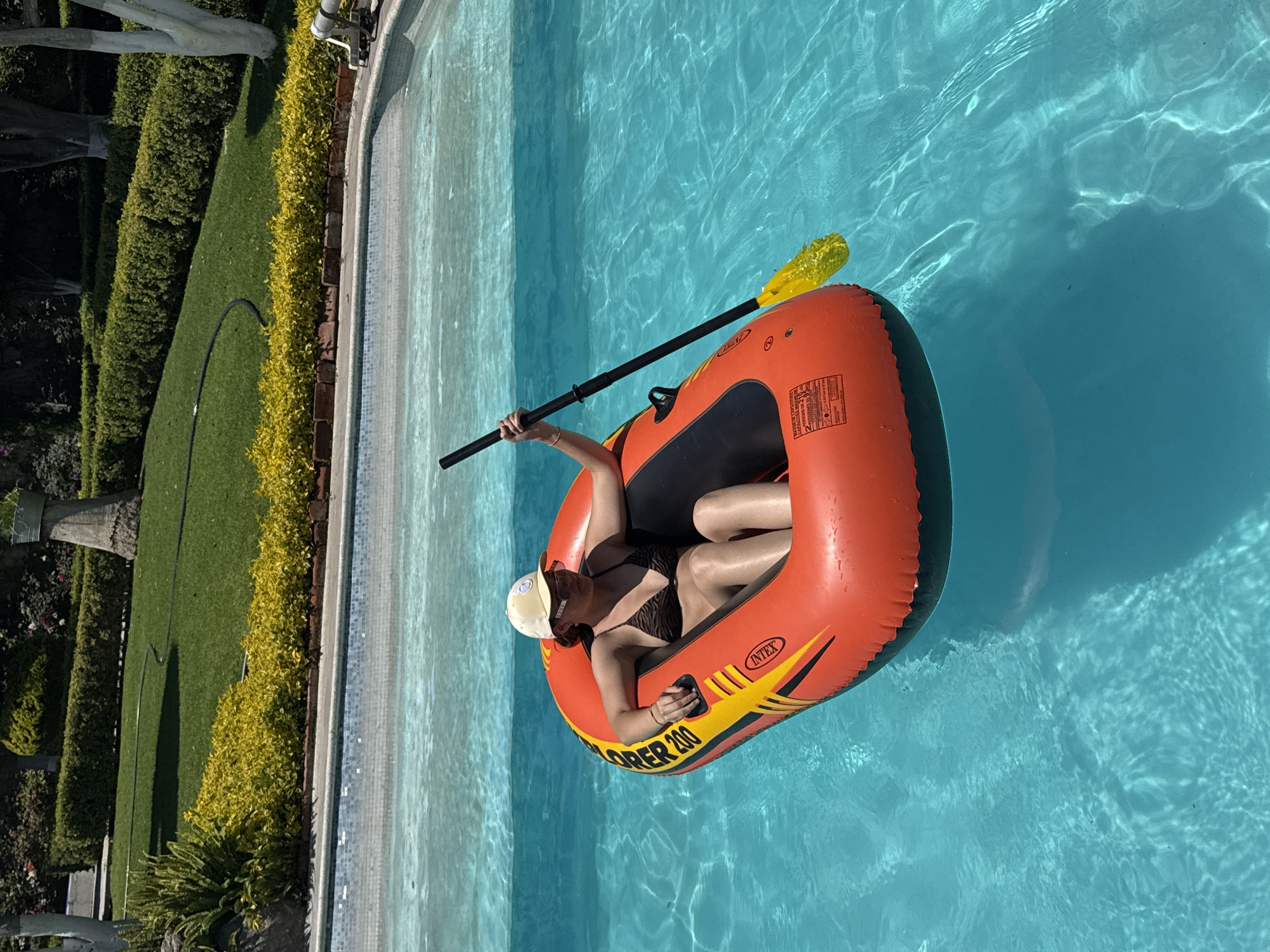A woman wearing a brown and black bikini and a white baseball cap is sitting in an orange inflatable raft in a swimming pool. She is holding a black paddle with a yellow blade in her right hand. The raft is labeled "Explorer 200" and "Intex". The pool is surrounded by a paved area with a low wall of hedges and grass. Beyond the hedges, there are trees and other greenery. The photo appears to have been taken in Puebla, Mexico, based on the architectural style and landscaping. The pool water is a clear blue.