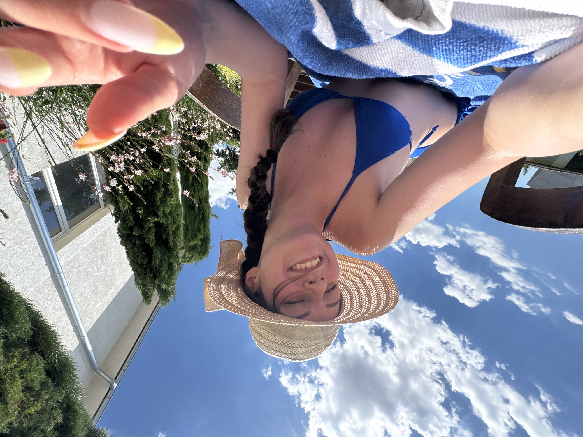 A young woman with a straw hat and blue bikini is lying on a towel outdoors on a sunny day. She is smiling with her eyes closed and her face tilted upwards towards the sky. The background shows a white building, green trees, and a blue sky with white clouds. The woman's left hand is in the foreground, with her fingernails painted in a yellow and pink French tip style. The scene appears to be a backyard or garden area, likely in a residential setting in the city of Bulgarovo, Bulgaria, given the architecture and vegetation.