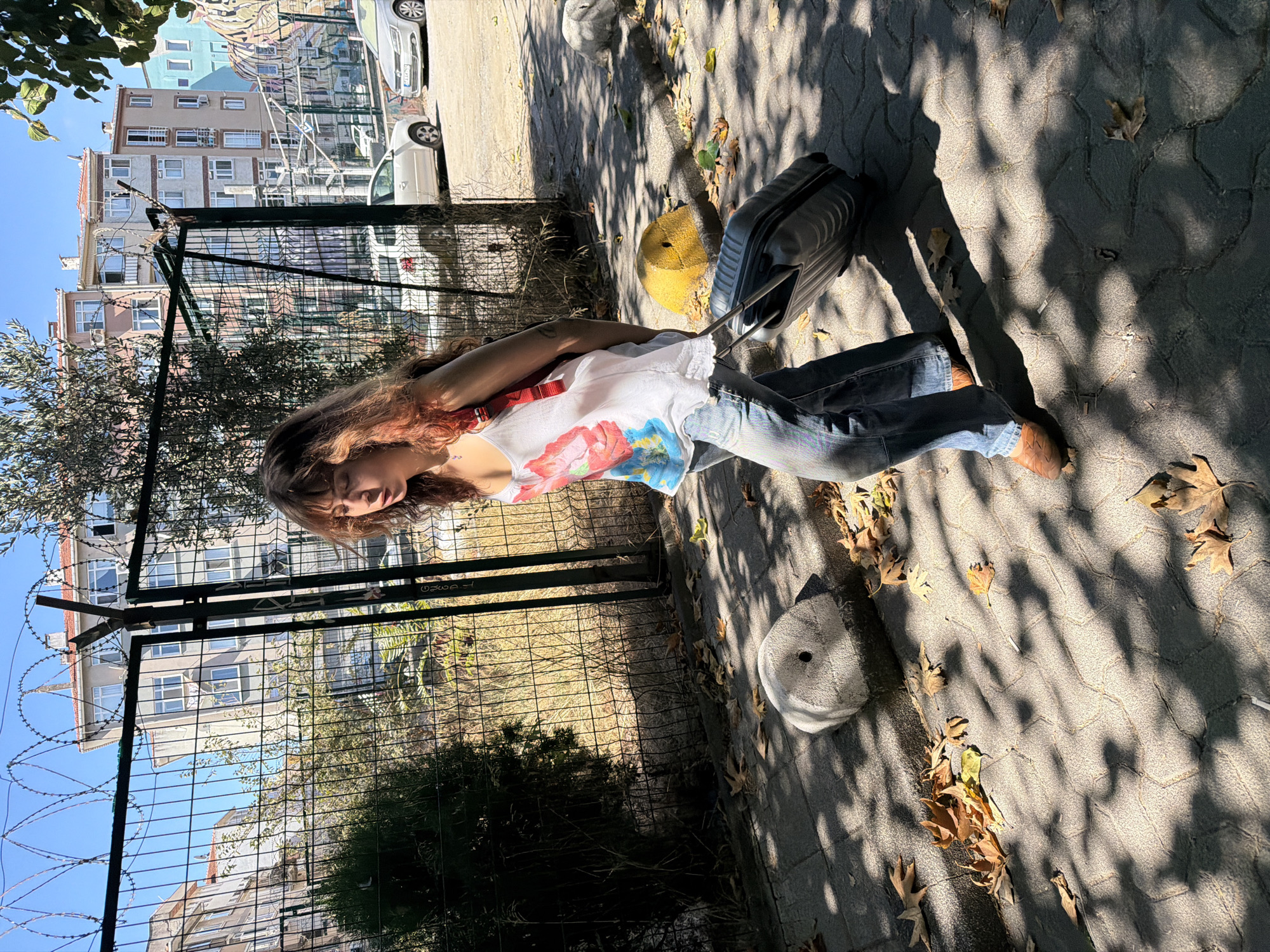 A young female with long, auburn hair walks on a cobblestone sidewalk, pulling a black rolling suitcase. She wears a white tank top with a floral print and blue jeans. The sun casts dappled shadows from overhead trees onto the sidewalk, where fallen leaves are scattered. Behind her is a metal fence, and beyond that, several apartment buildings are visible. In the background, cars are parked on the street. The scene appears to be in a residential area of Kadıköy, Turkey, given the architectural style of the buildings and the general urban setting.