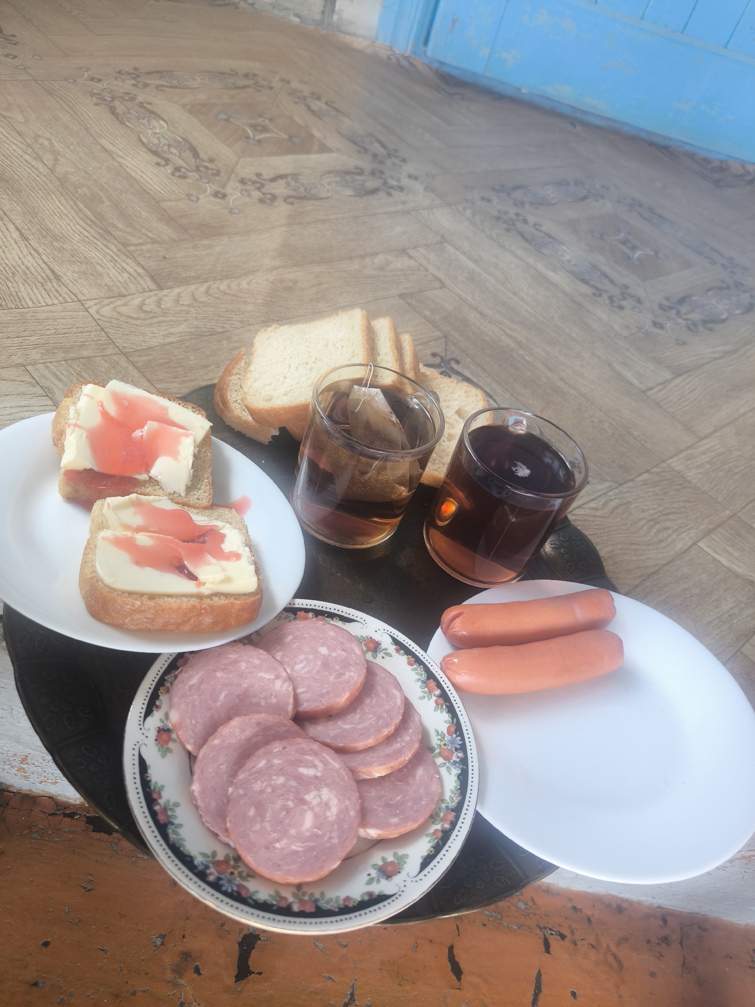 The image captures an overhead view of a meal laid out on a dark, patterned surface, likely a tray or small table, positioned over a weathered orange-brown surface. The background consists of a light brown, patterned wooden or laminate floor, and a distressed, light blue wooden structure in the upper right.
The meal comprises several components:
1. **Plates and Food:**
* A white, rectangular plate in the mid-left holds two slices of bread, each topped with a thick slab of pale yellow butter and a zigzag drizzle of red syrup or jam.
* A round, white plate with a dark blue rim adorned with a floral pattern, located in the lower left quadrant, contains approximately ten circular slices of light pink cured sausage, possibly salami.
* A plain white, rectangular plate in the lower right displays two orange-pink frankfurter-style sausages.
* Behind the buttered bread and glasses, several plain slices of white bread are visible.
2. **Beverages:**
* Two clear glass mugs, positioned centrally, contain a dark brown liquid, consistent with tea. A teabag is visible steeping in the left mug.
3. **Surfaces:**
* The primary dark surface holding the food items has a subtle embossed pattern and appears reflective.
* Below this, an older, possibly wooden or concrete, surface with extensive peeling orange-brown paint and visible cracks provides a weathered texture.
* The dominant background is a light brown floor featuring a repeating pattern of dark grey/brown ornate swirls and geometric shapes.
* In the upper right, a section of a light blue wooden structure, exhibiting paint wear and texture, is visible.
There are no individuals present in the image. The setting suggests an indoor or sheltered outdoor environment in the city of Unknown, Unknown.