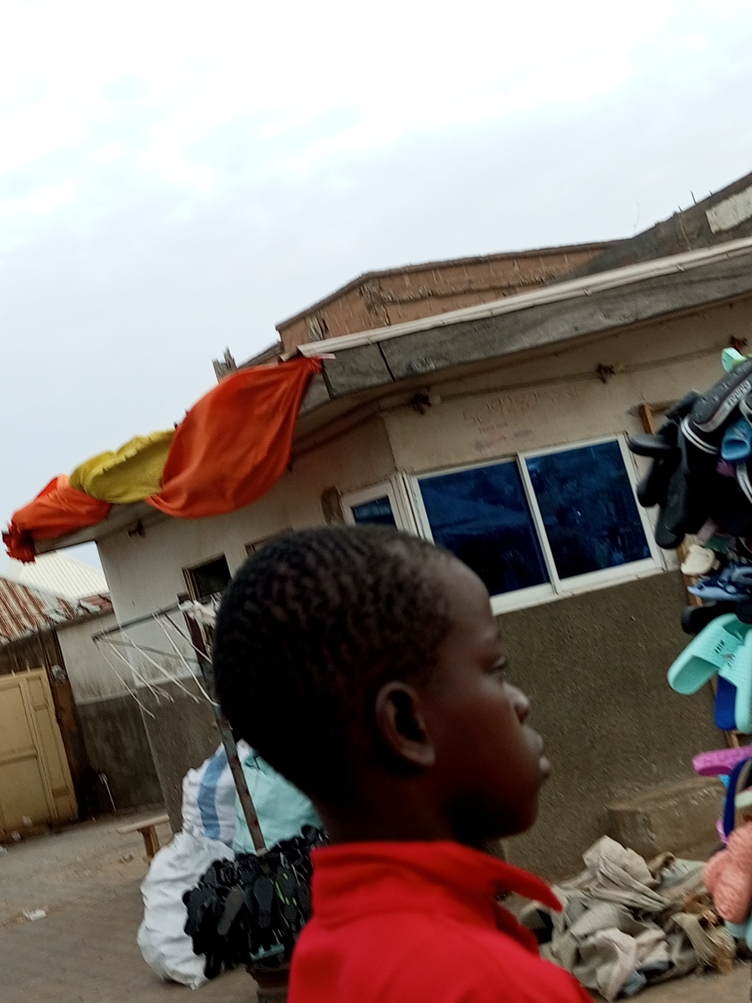 A young Black boy wearing a red shirt stands in profile, looking to his right. He is in an outdoor market setting. Behind him, a building with blue windows is visible, and a vibrant orange and yellow awning or fabric stretches across the top. Several pairs of footwear, including sandals and flip-flops, are displayed on a rack to the right of the boy. In the foreground and to the left, sacks of goods and a pile of black shoes are present. The sky is overcast. The scene appears to be in Tempure, Nigeria.
