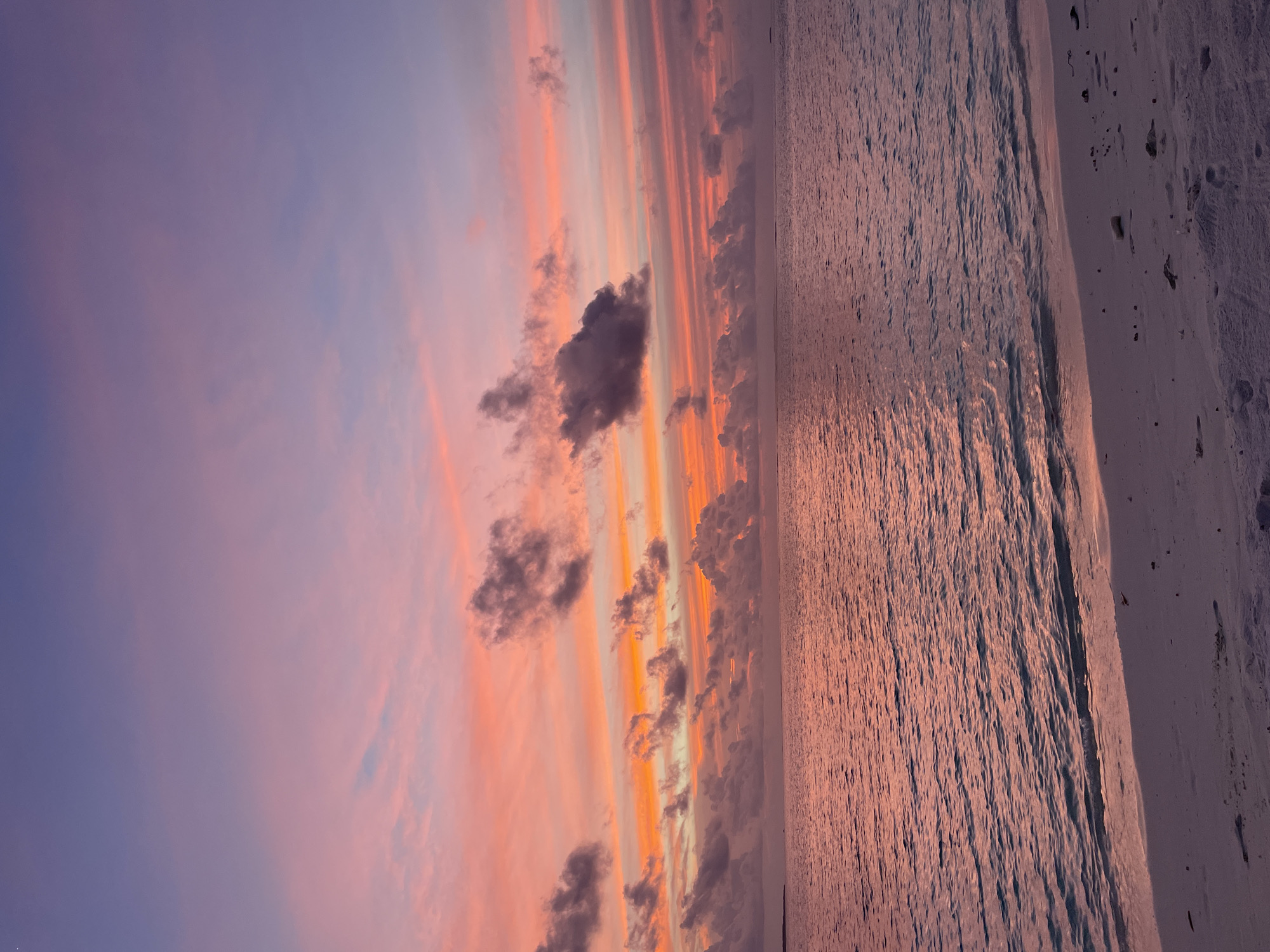 The image depicts a serene beach scene at sunset. The sky is painted with vibrant hues of purple, pink, and orange, with wispy clouds scattered across the horizon. The sun, not directly visible, casts a warm glow that reflects off the calm ocean waters. The water's surface is textured with gentle ripples, creating a shimmering effect. To the right, a sandy beach stretches into the foreground, showing imprints of footprints, suggesting recent activity. The overall mood is peaceful and tranquil, evoking the beauty of a coastal evening. The time of day is clearly sunset, indicated by the warm, diffused light and the colorful sky. There are no visible objects or text in the image.