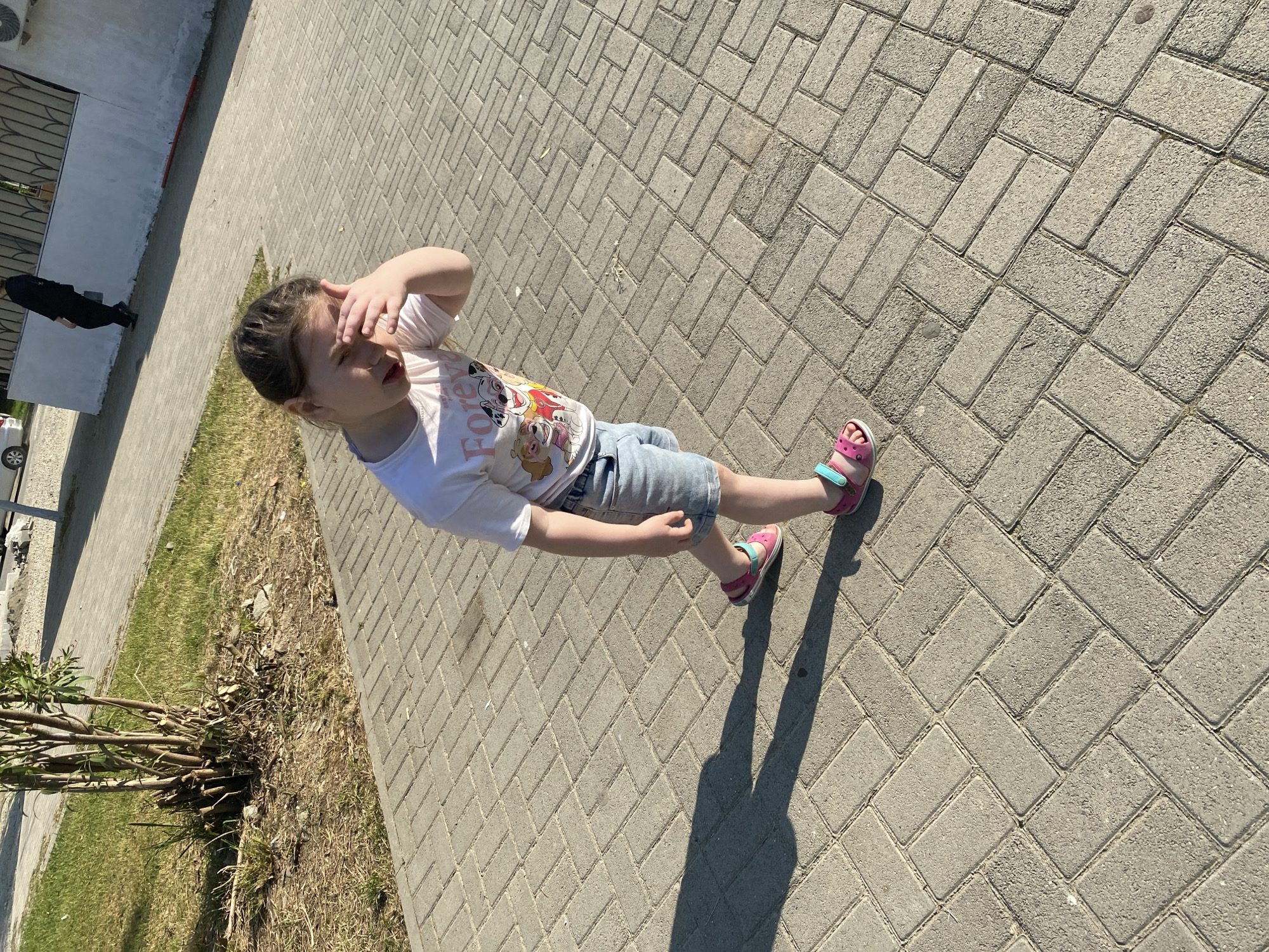 A young girl wearing a white t-shirt with a Paw Patrol graphic and denim shorts stands on a herringbone-patterned brick sidewalk in a city. She has her hand over her eyes, as if shielding them from the sun. Her pink sandals are visible. In the background, a person in dark clothing walks away from the camera, and a white wall with a black railing stands behind them. To the left of the sidewalk, there is a patch of dry grass and a small, bush-like plant. The lighting suggests it is a sunny day.