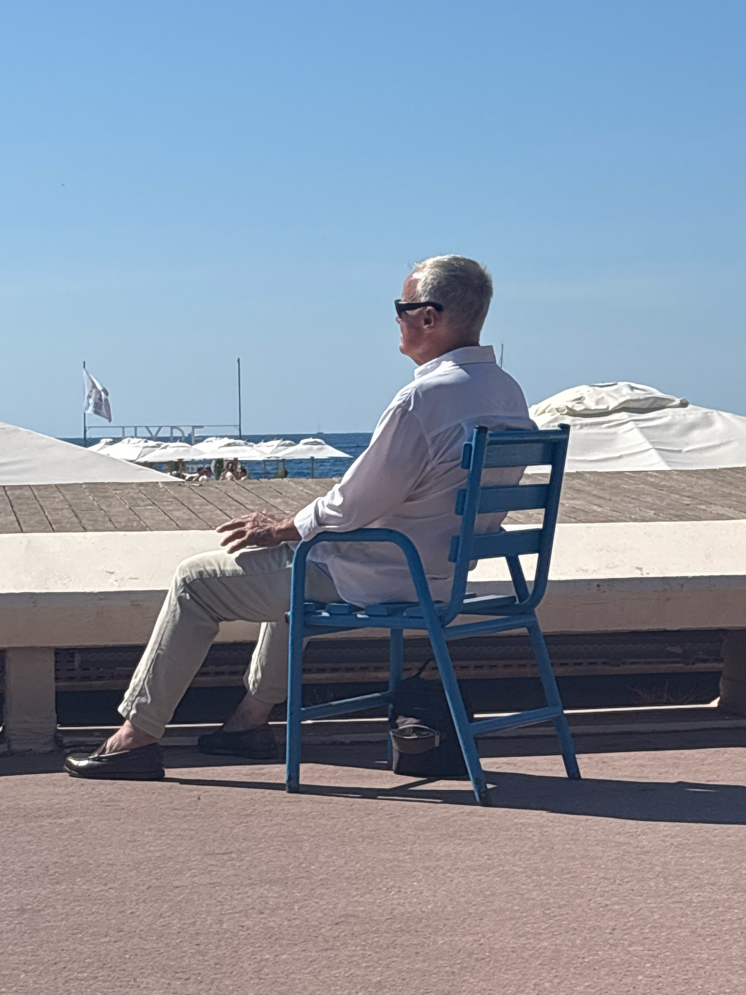 The image depicts a man sitting alone on a bright blue chair on a sunny day, facing away from the camera. He has light-colored hair and is wearing sunglasses, a white collared shirt, and light-colored pants. His left hand rests on his lap. The chair is positioned on a paved surface, with a wooden deck and beach umbrellas visible in the background. Beyond the deck is the ocean and a clear blue sky. A white flag with a logo is visible on a pole in the distance. The overall setting appears to be a beach or waterfront area.