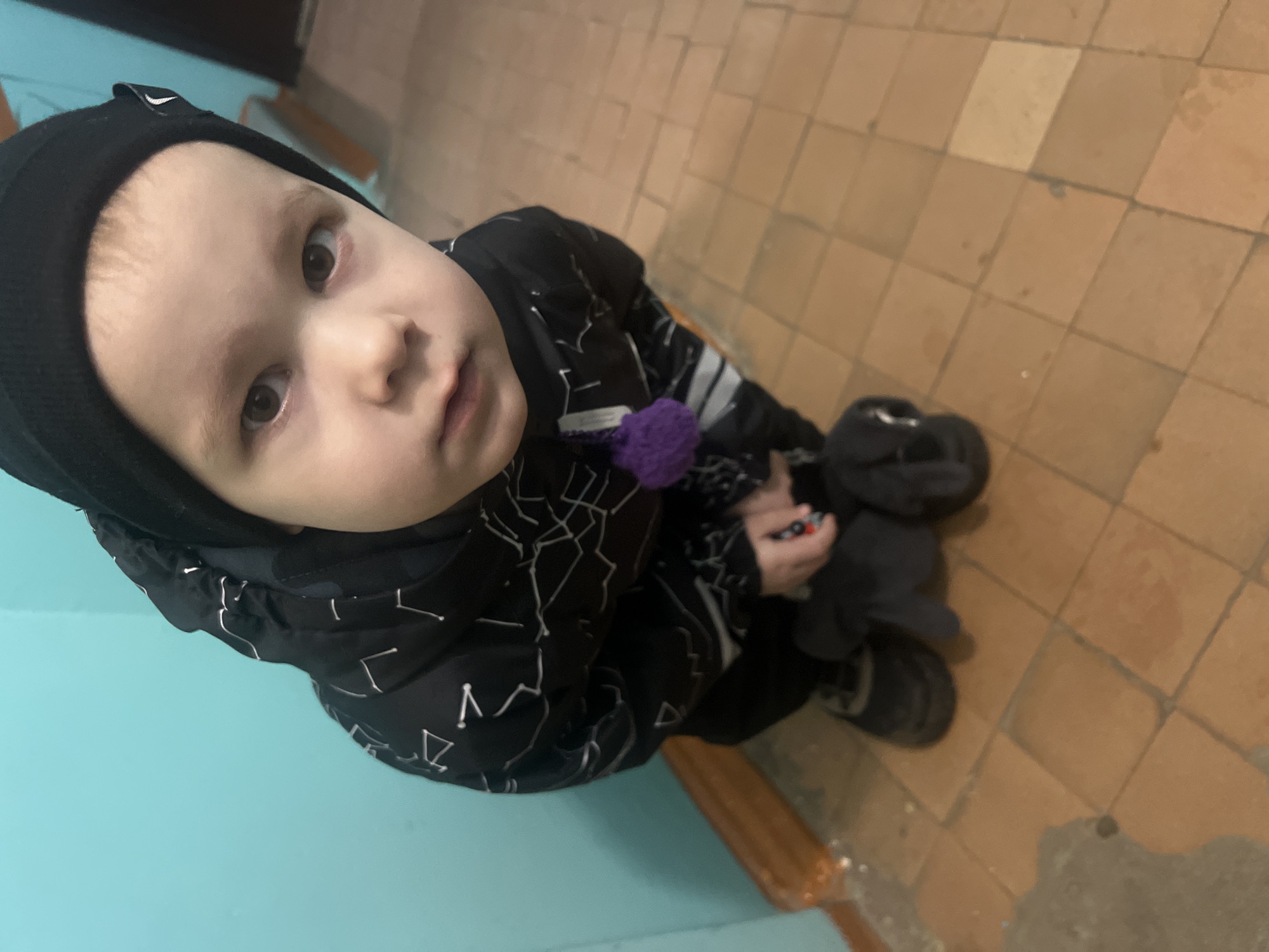 A young child, approximately 2-4 years old, with light skin and dark eyes, is captured in a high-angle, eye-level shot, looking directly at the viewer. The child wears a black knitted beanie cap and a black hooded jacket adorned with white abstract or constellation-like patterns. A small purple knitted pom-pom is attached to the jacket's left side. The child is crouched or kneeling, wearing black trousers and dark, possibly black, shoes. Their right hand appears to be grasping a small, dark object, possibly a toy, with a red and black detail visible near the fingers. The left hand also holds a dark, soft object. The child is positioned against a bright turquoise-colored wall, which shows some wear and a dark wooden baseboard at its bottom edge. The floor is composed of light brown or reddish-brown square tiles with visible grout lines and some surface discoloration or marks. In the upper right background, a dark vertical element, possibly a door frame or cabinet edge, is discernible. No specific visual markers identifying the exact location within Chaikovsky, Russia, are present in the image.