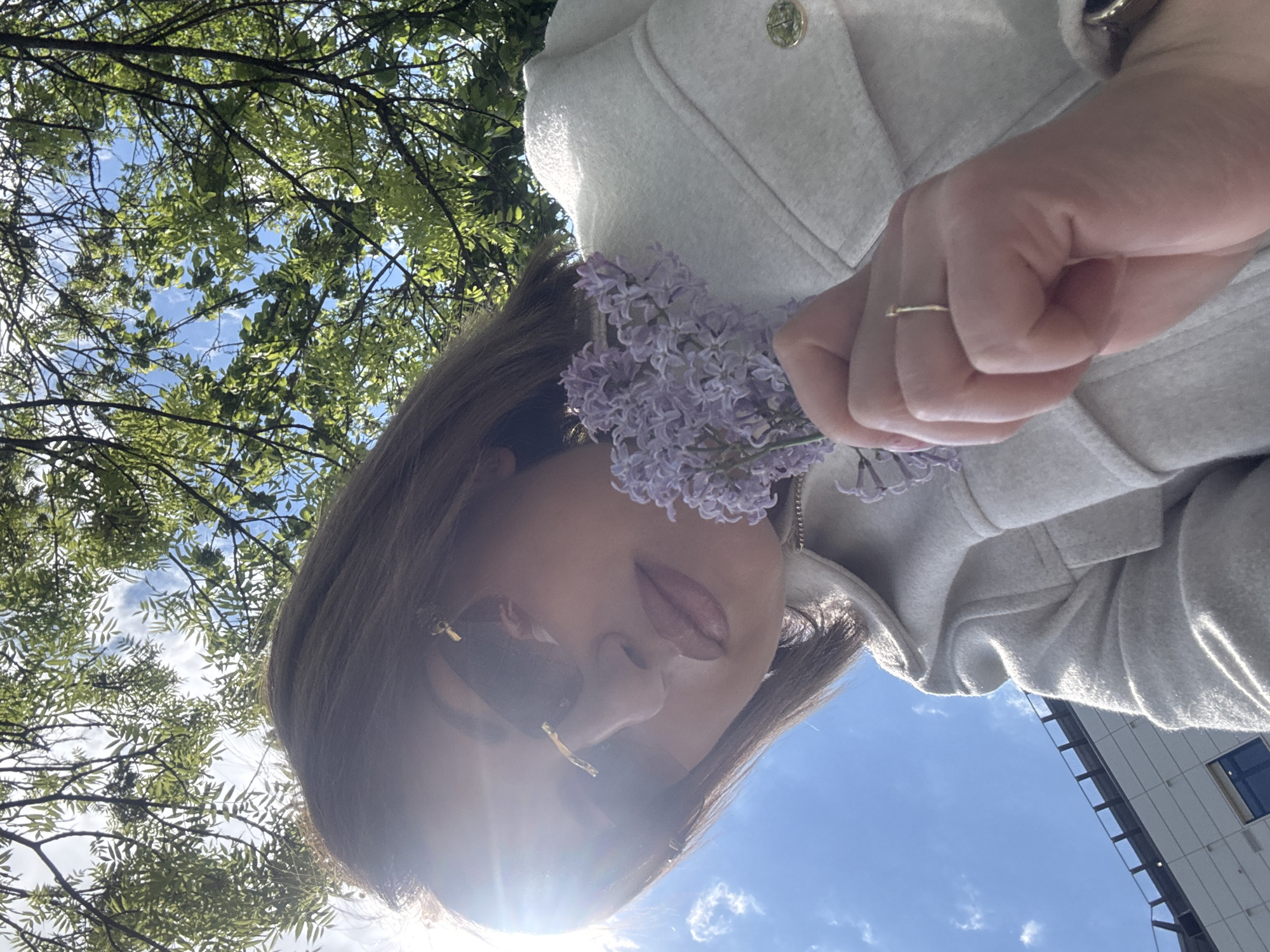 A low-angle shot depicts a woman holding a bouquet of purple flowers. She is wearing sunglasses and a light-colored jacket. The background includes tree branches with green leaves against a blue sky, suggesting an outdoor setting. A building is partially visible in the background. Given the provided context, the location is assumed to be in Sofia, Bulgaria, though specific landmarks are not visible to confirm this.
