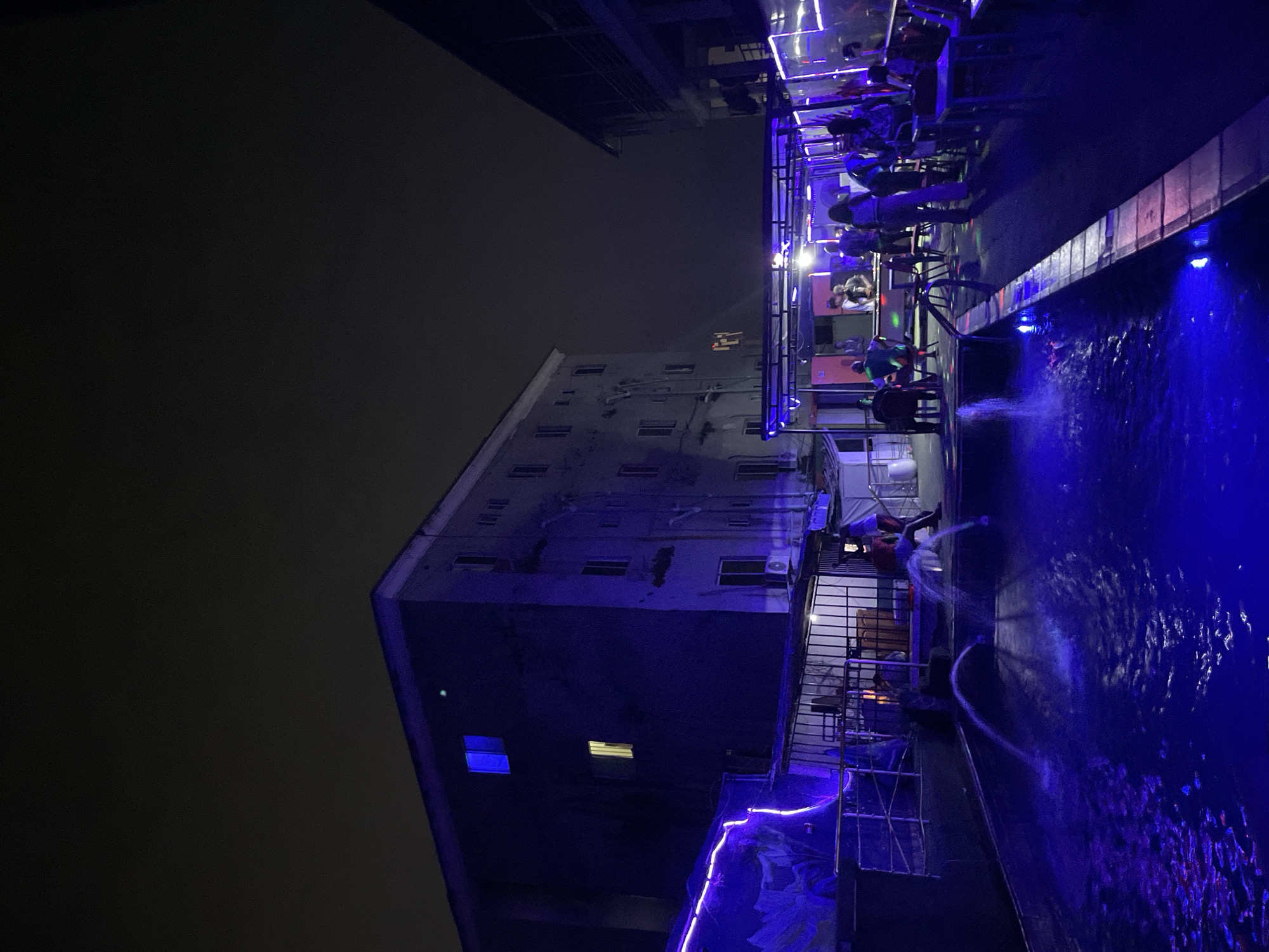 This night-time photograph, reportedly taken in Ado, Nigeria, depicts an illuminated outdoor rooftop or elevated venue, possibly a bar or restaurant, positioned next to a vibrant swimming pool. The scene is bathed in a predominant blue light, originating from underwater pool lighting and decorative neon strips that outline the structures.
On the right side of the image, several people are visible, mostly seated at tables in a covered seating area, illuminated by spotlights. They appear to be socializing or relaxing. One individual stands near the pool's edge, where multiple water jets create dynamic white splashes and streaks against the deep blue water. The pool's surface exhibits ripples, reflecting the ambient light.
To the left, a dark, multi-story building rises against an equally dark, featureless night sky. Its exterior appears greyish-blue under the ambient lighting, with several windows, some dark and others emitting warm light from inside. Metal railings and other structural elements delineate the boundaries of the seating and poolside areas. The setting conveys an atmosphere of modern evening entertainment, characterized by its distinctive blue lighting and the lively water features. No discernible text is visible within the image.