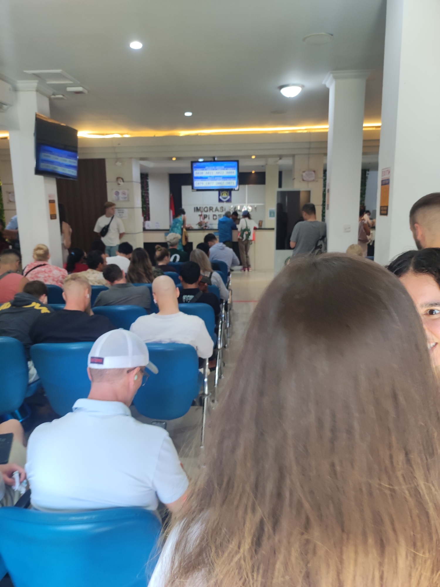 The image shows a waiting area, potentially at an immigration office, filled with seated people. The location is suggested to be in Denpasar, Indonesia. Rows of blue plastic chairs are occupied by individuals of various ages and appearances. At the far end of the room, a service counter is visible, with signage indicating "IMIGRASI TPI DE" (Immigration TPI DE). Display screens are mounted on the wall above the counter. The room appears well-lit, with white walls and pillars. The people in the waiting area seem to be waiting patiently. The perspective is from behind a person with long brown hair, partially obscuring the view, with a glimpse of another person to the right side of the frame.