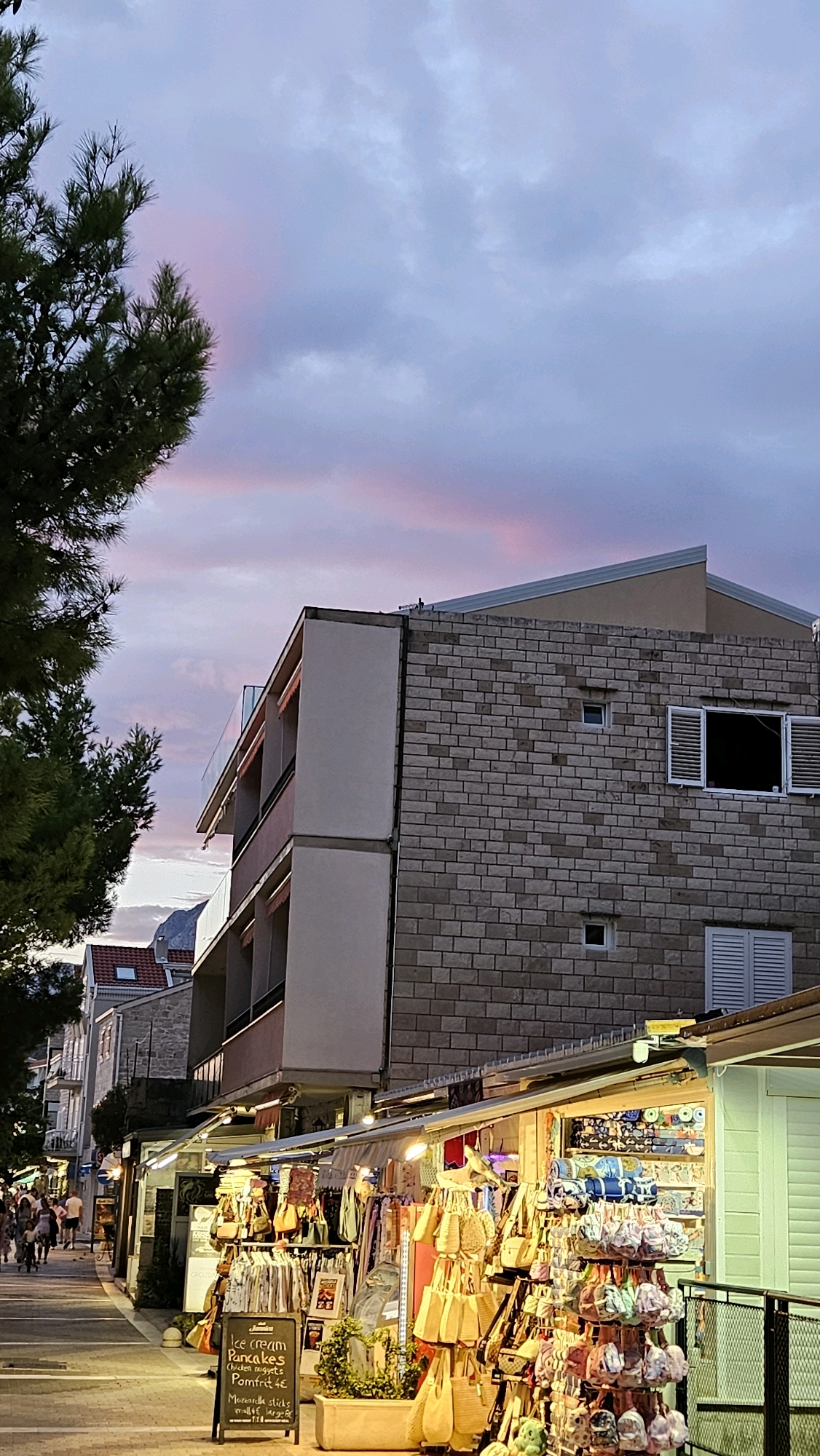 This is a vertical, full-frame, eye-level shot of a street scene at dusk in Općina Tučepi, Croatia. The sky displays a dramatic twilight with streaks of pink and purple clouds against a blue-gray background. A large pine tree with dark green needles dominates the upper left portion of the frame, casting a shadow on the scene below.
In the midground, a multi-story building with a facade of light brown brick is visible. The building features several windows, some with white shutters, and balconies with glass railings. To the left of the brick building, a lighter-colored building with multiple floors and balconies can be seen, with awnings extending over some of the balconies.
The foreground is occupied by a bustling street lined with shops and market stalls. The stalls are illuminated by warm, artificial lights and display a variety of goods, including clothing, handbags, and souvenirs. Several racks of clothing and bags are prominently featured. A black chalkboard sign in front of one of the stalls lists food items like "ice cream," "pancakes," "chicken nuggets," and "pomfrit 4€". A small potted plant sits in a planter box near the sign.
On the left side of the street, a paved walkway is visible with several people walking in the distance. The overall impression is a vibrant and lively atmosphere as evening descends.