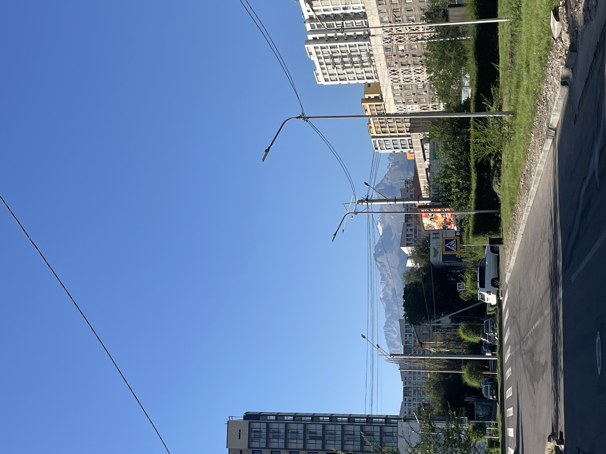 This is an eye-level, vertical, full shot of a city street with mountains in the background, taken in Almaty, Kazakhstan. The sky is clear and blue, with a power line running diagonally from the upper left to the middle of the frame. In the foreground, a street with white crosswalks is visible, with a gray asphalt surface and a concrete curb to the right. To the left of the street is a sidewalk and a large, modern, light-colored apartment building with many windows. In the middle ground, several multi-story buildings with different architectural styles and colors are visible, some appearing to be residential or office buildings. A white SUV is parked on the right side of the street, and other vehicles are parked on the left. Streetlights are present along the road, and lush green trees and bushes line the median and the sides of the road. In the background, majestic, snow-capped mountains rise above the city buildings, forming a striking contrast with the urban landscape. The overall scene suggests a bright, sunny day in a city known for its proximity to natural mountain scenery.