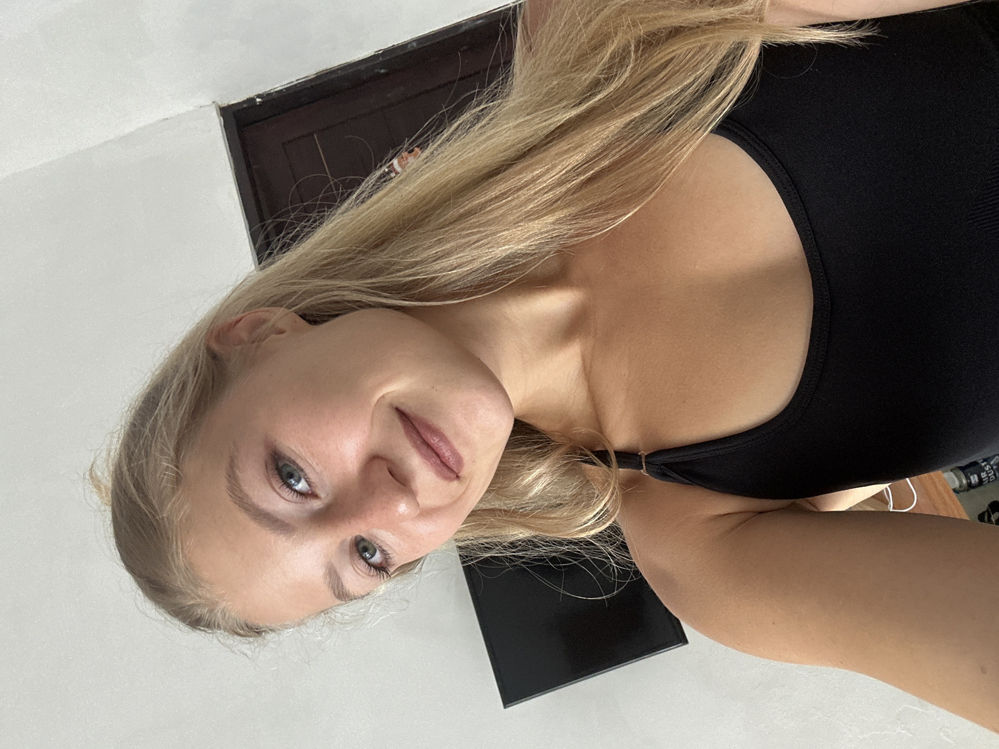 The image, taken in Kuta Selatan, Indonesia, features a young woman in a close-up selfie, oriented diagonally, looking directly at the camera with a gentle, relaxed smile. She has long, straight blonde hair that cascades around her, bright blue or grey eyes, and fair skin with a slight tan. She is wearing a fitted black tank top or sports bra. Her lips are lightly tinted, and her eyebrows are well-defined.
The setting appears to be indoors with a minimalist background. A white wall dominates the upper left, while a dark wooden door or door frame is visible in the upper right corner, creating a stark contrast. A dark, rectangular object, possibly an electronic device or furniture, is partially visible on a white surface to her lower left. The lighting is bright and even, illuminating her face and the immediate surroundings without harsh shadows. No discernible text is visible within the frame.