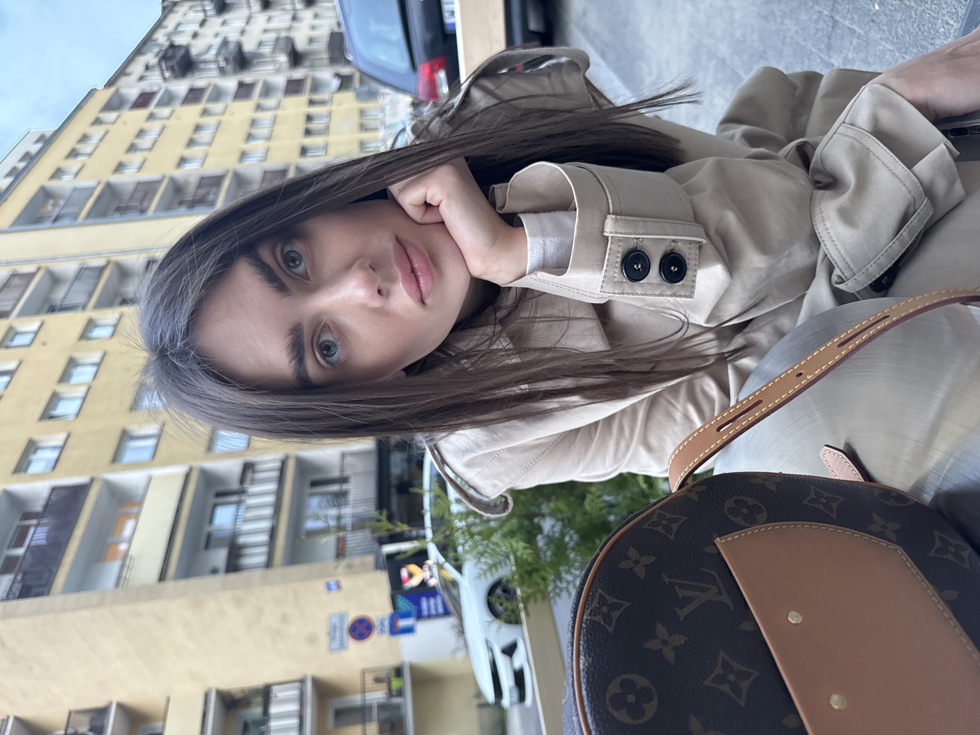 A woman with long brown hair is sitting outdoors, possibly at a cafe, in Warsaw, Poland. She is wearing a light beige trench coat with a white top underneath. Her left hand is resting on her cheek. A Louis Vuitton bag is partially visible in the foreground. Behind her, there is a street scene with a parked car and a building that appears to be an apartment complex, identifiable with elements of typical Warsaw architecture and street signs visible. The image has been taken at a tilted angle.
