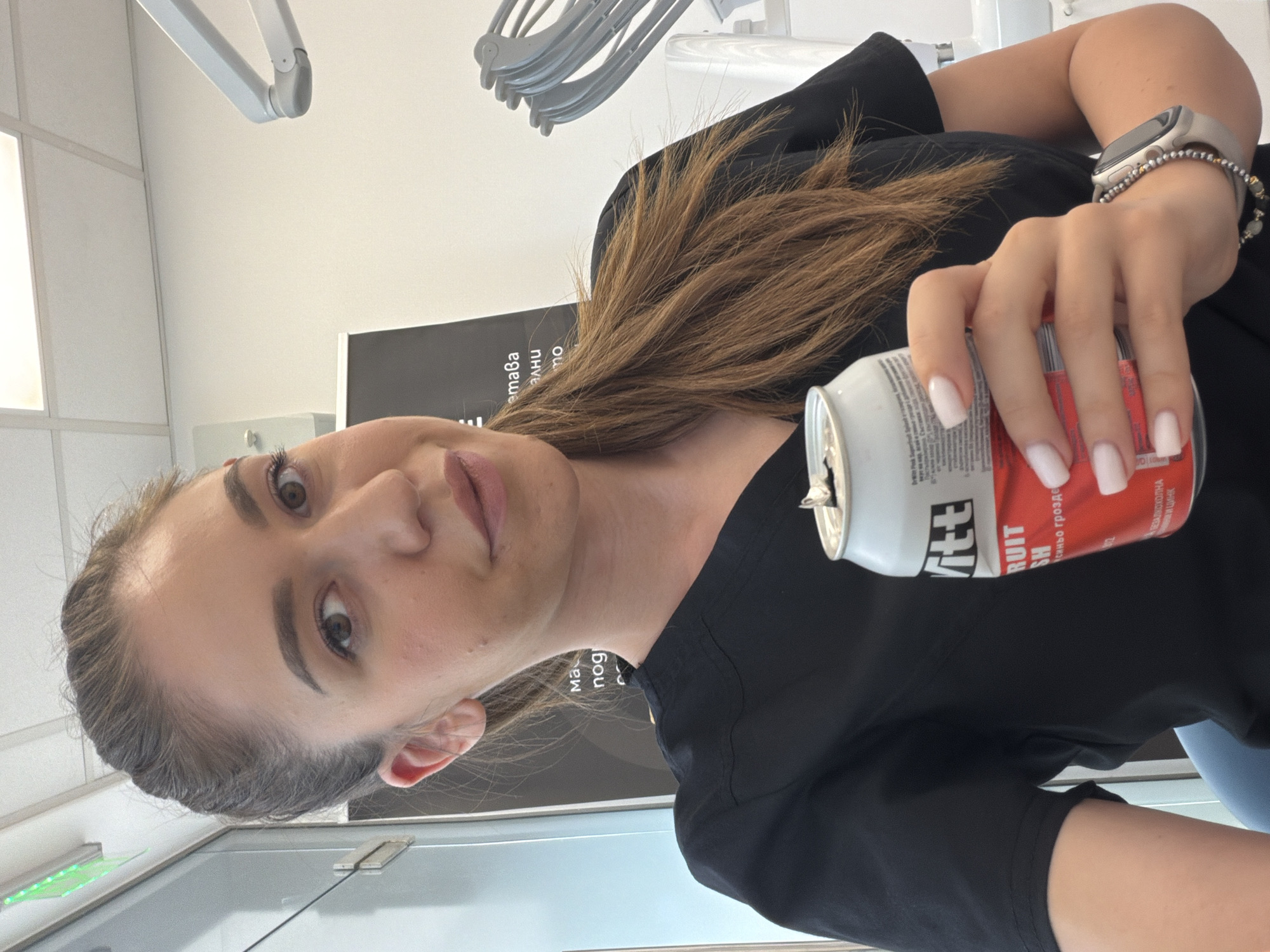The media file is a vertical, medium close-up selfie of a young woman in what appears to be a dental office. She is wearing a black scrub top and a smartwatch on her left wrist. She holds a red and white can of "Fruity Rush" beverage in her right hand, with her fingers curled around it. Her hair is pulled back in a ponytail.
The woman has light skin and is looking directly at the camera with an engaged expression. Her makeup is subtle, with emphasized eyebrows and mascara. Her fingernails are painted a clean, bright white. The background shows part of a dental examination light, a wall with some sort of grid paneling, and a black banner with white text, some of which is visible. The text on the banner appears to be in Cyrillic script and includes the words "тава" and "нощ".
The scene is indoors and well-lit, suggesting daytime. The presence of medical equipment and the banner indicate a professional setting, likely a dental clinic. The woman's relaxed posture and direct gaze suggest a casual moment captured during her workday. There are no indications of weather or specific time of day beyond the indoor lighting.