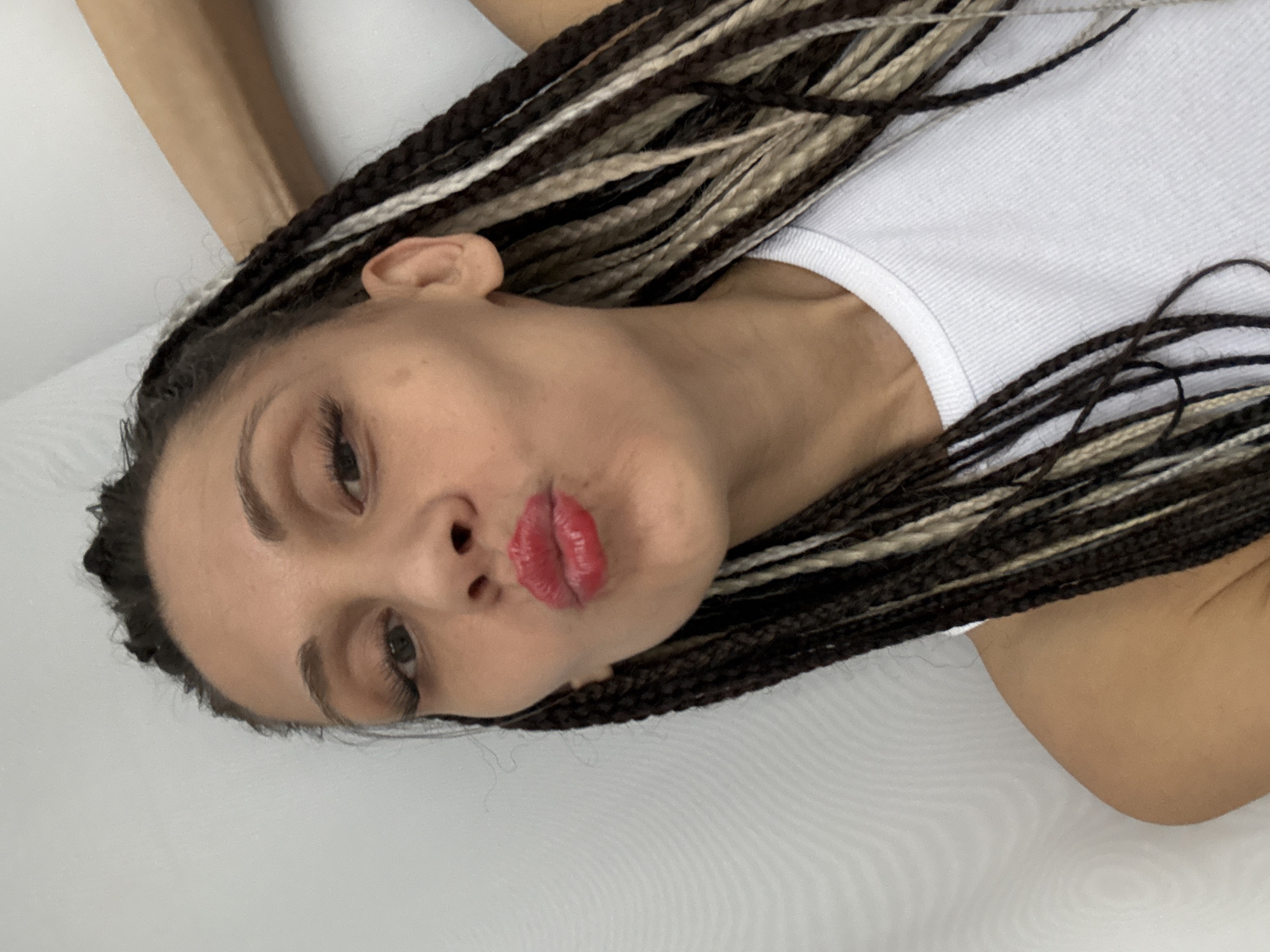 A woman with braided hair, a white tank top, and red lipstick is positioned against a white background. The location and city are unknown.