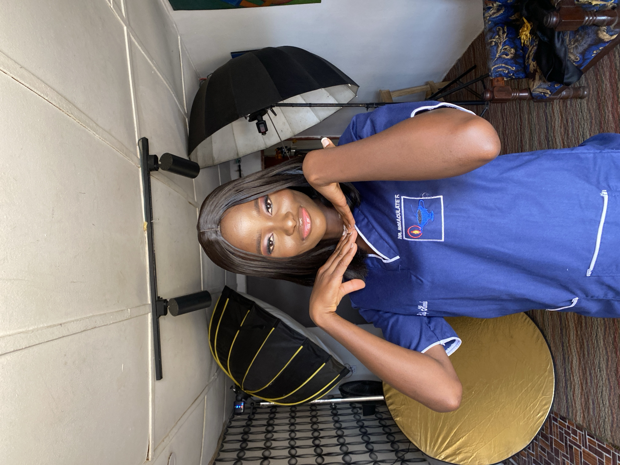 A young woman with dark skin and long, straight dark hair is featured in a portrait photograph, taken indoors, likely in Vom, Nigeria. She is looking directly at the camera with a gentle smile and soft, glossy lips, her eyes accentuated with makeup. She wears a navy blue top with white trim, resembling a uniform or scrubs, which has an embroidered logo on the left chest. The text "NR. IMMACULATE R" is clearly visible on the logo. Her hands are elegantly posed near her face, palms slightly open.
The setting appears to be a photography studio or a dedicated space for photoshoots. Various pieces of professional photography equipment are visible: a large black umbrella or softbox positioned behind her to her left, a smaller black softbox with yellow accents placed closer to the foreground on her right, and a prominent gold circular reflector on the floor beside her. These items indicate controlled lighting for the portrait.
The background includes a light-colored paneled wall on one side and a multi-toned, patterned rug on the floor. A wooden chair with blue and gold patterned upholstery is partially visible behind her to her right. The overall atmosphere suggests a professional and deliberate photoshoot.
