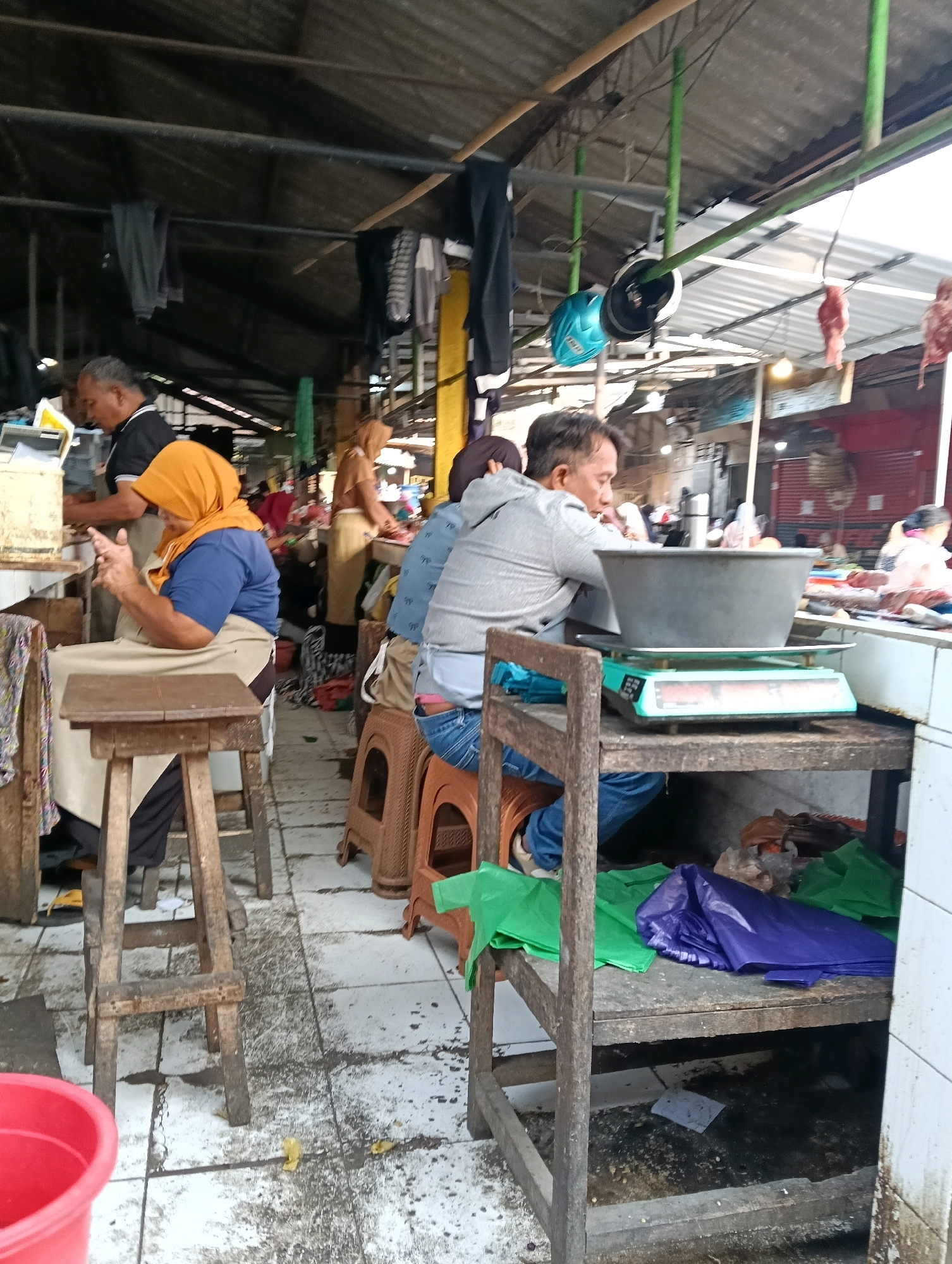 This is a mid-range shot of a market scene in Magelang, Indonesia. In the foreground, a woman wearing a blue shirt and an orange headscarf sits on a wooden stool next to a wooden counter. Her hands are clasped, and she appears to be engaged in conversation or waiting. To her right, a man in a grey hooded sweatshirt and blue jeans sits on a stool, looking down at a scale with a large metal bowl on it. Behind the woman, a man in a black shirt is visible behind a counter, possibly working.
The environment appears to be an open-air market with a corrugated metal roof supported by wooden poles. Various items are hanging from the ceiling, including what appear to be articles of clothing and some raw meat. Stalls are set up with counters displaying goods, and plastic bags of various colors (green, blue, purple) are stacked on shelves and surfaces. The floor is tiled and appears somewhat wet and dirty, with some debris scattered around. The overall impression is a busy, somewhat unkempt, but functional marketplace.