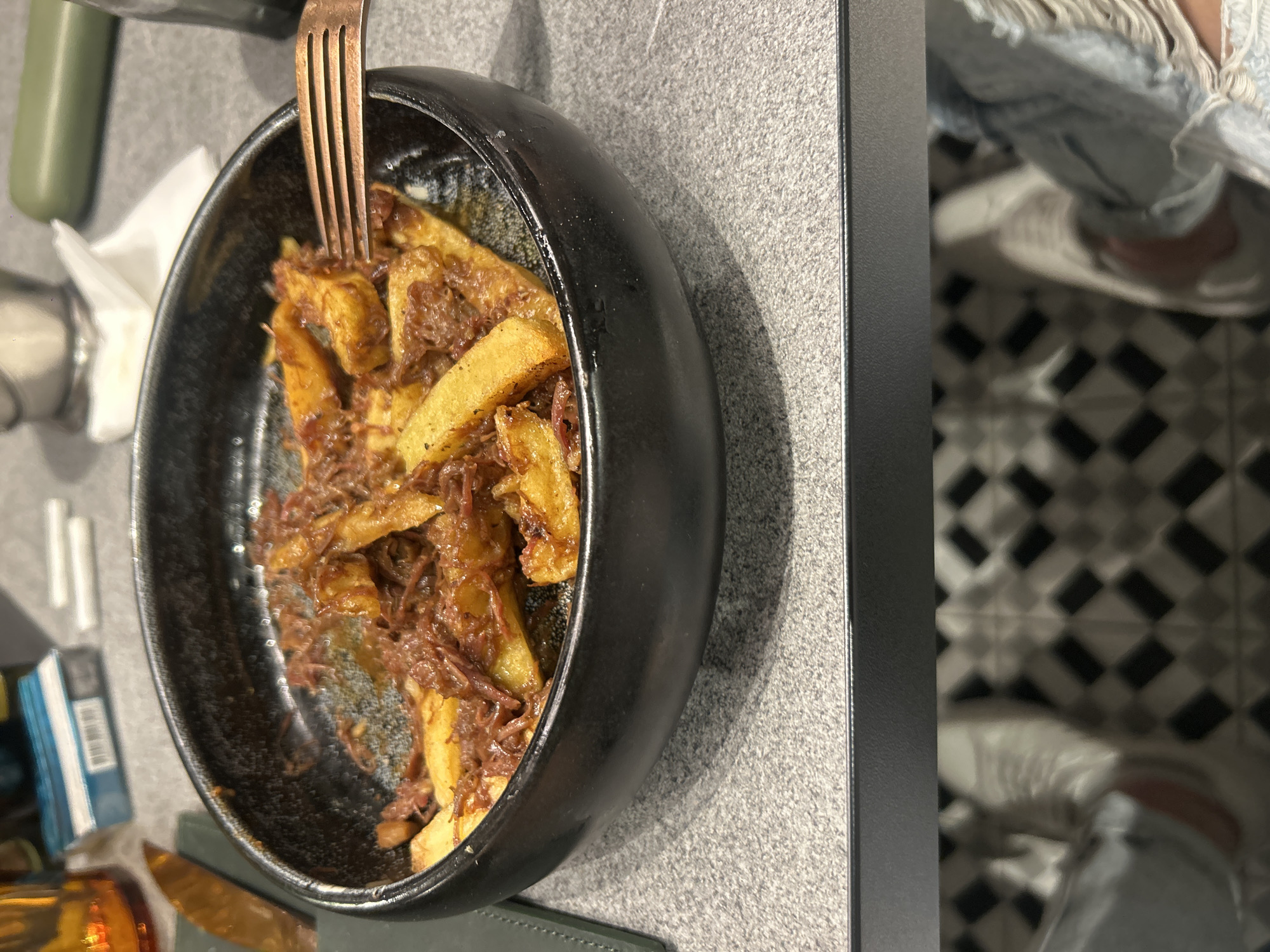 A high-angle shot shows a partially eaten plate of food. The food consists of french fries covered in a brown, meaty sauce, served in a dark-colored, speckled bowl. A copper-colored fork is stuck into the food. The bowl sits on a gray countertop next to a black partition. To the left of the bowl are partial views of other items, including a green container, napkins, a salt shaker, and what appears to be a pack of cigarettes. The bottom of the frame shows the edge of a green place mat and an amber-colored glass. To the right, reflecting on the black partition, is a glimpse of a black and white tiled floor and the legs and feet of a person wearing jeans and white sneakers. Given the types of foods and general scene this image might be from a restaurant or cafe. Without additional context, it's difficult to confirm if it is located in Plovdiv, Bulgaria.