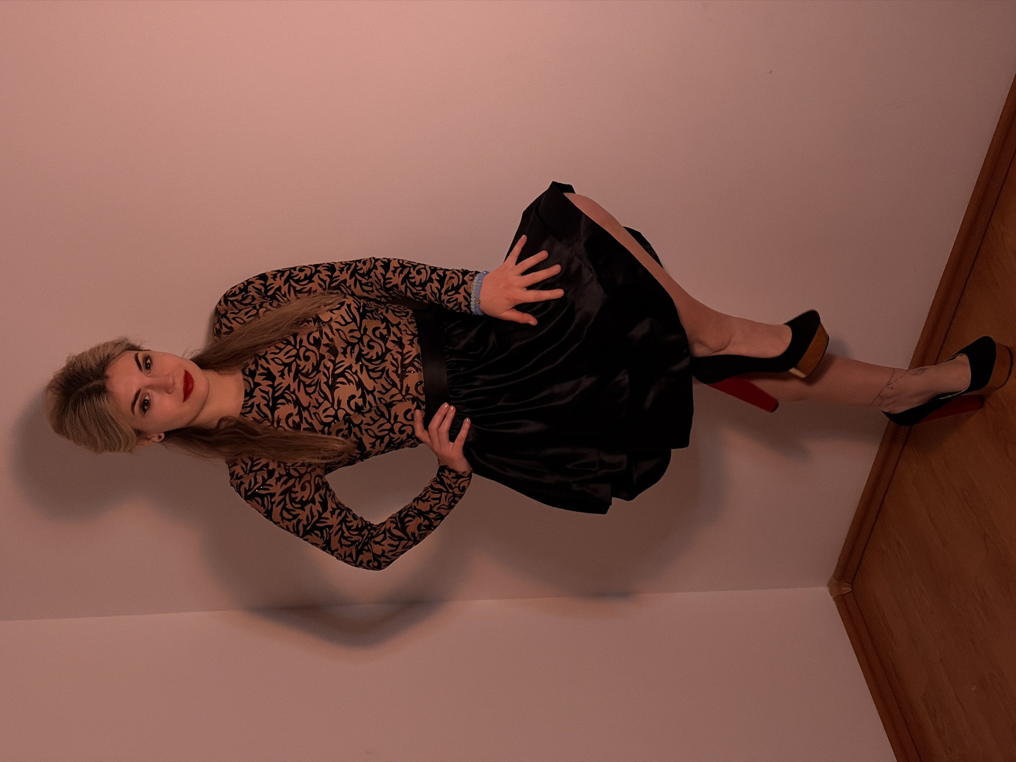 The image displays a full-length shot of a woman posing indoors, rotated ninety degrees clockwise. She is positioned in the corner of a room, with plain light pink or peach-colored walls forming the background, and a wooden floor or paneling visible along the right side of the frame.
The woman has long, straight blonde or light brown hair, styled with some volume around her face. She is looking directly at the camera with a subtle smile. Her makeup includes prominent red lipstick and defined eye makeup. She is wearing a long-sleeved top with a black and gold/tan patterned design that appears to be intricate and perhaps shimmery or sequined. The top features a high neckline. Below the top, she wears a black, high-waisted skirt that has a satin or silky texture, appearing to be full and pleated or gathered, ending around her knees.
Her pose is dynamic; she is leaning back slightly, with her right hand resting on her hip and her left hand gently placed on her upper left thigh or the skirt. Her legs are bent, and she is wearing black high-heeled shoes with a chunky wooden platform and a distinctive red sole, suggesting a designer brand. A light blue beaded bracelet is visible on her left wrist.
The lighting in the room appears to be warm and artificial, casting soft shadows on the walls and highlighting the textures of her clothing. No text is visible in the image. The overall impression is a posed portrait, likely taken in the evening or indoors with controlled lighting.