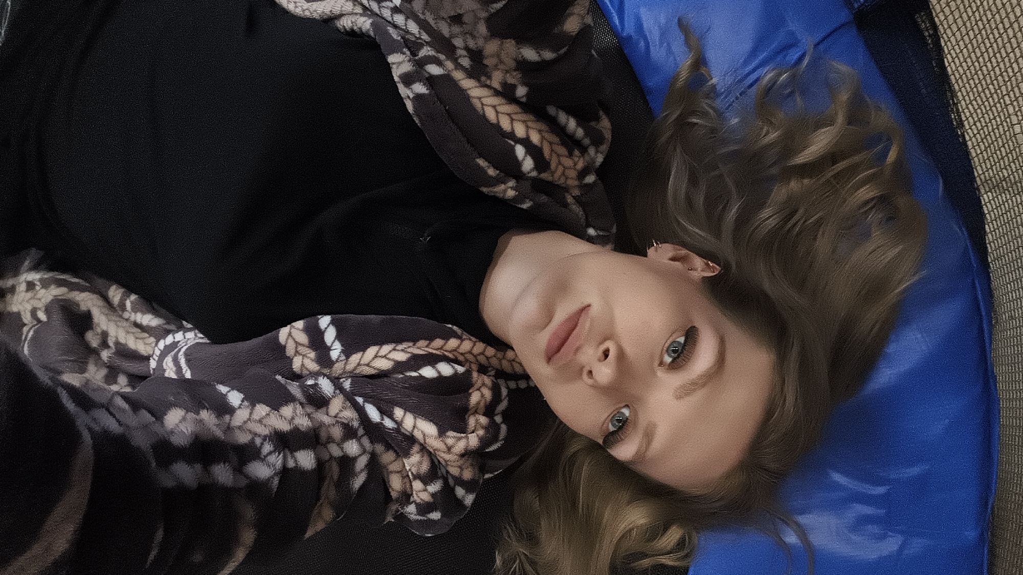 A woman lies on her back on a blue surface with curled hair. She wears a black top and a patterned scarf or shawl. The background suggests an indoor setting, possibly a trampoline. The location is unspecified and cannot be inferred from the image.