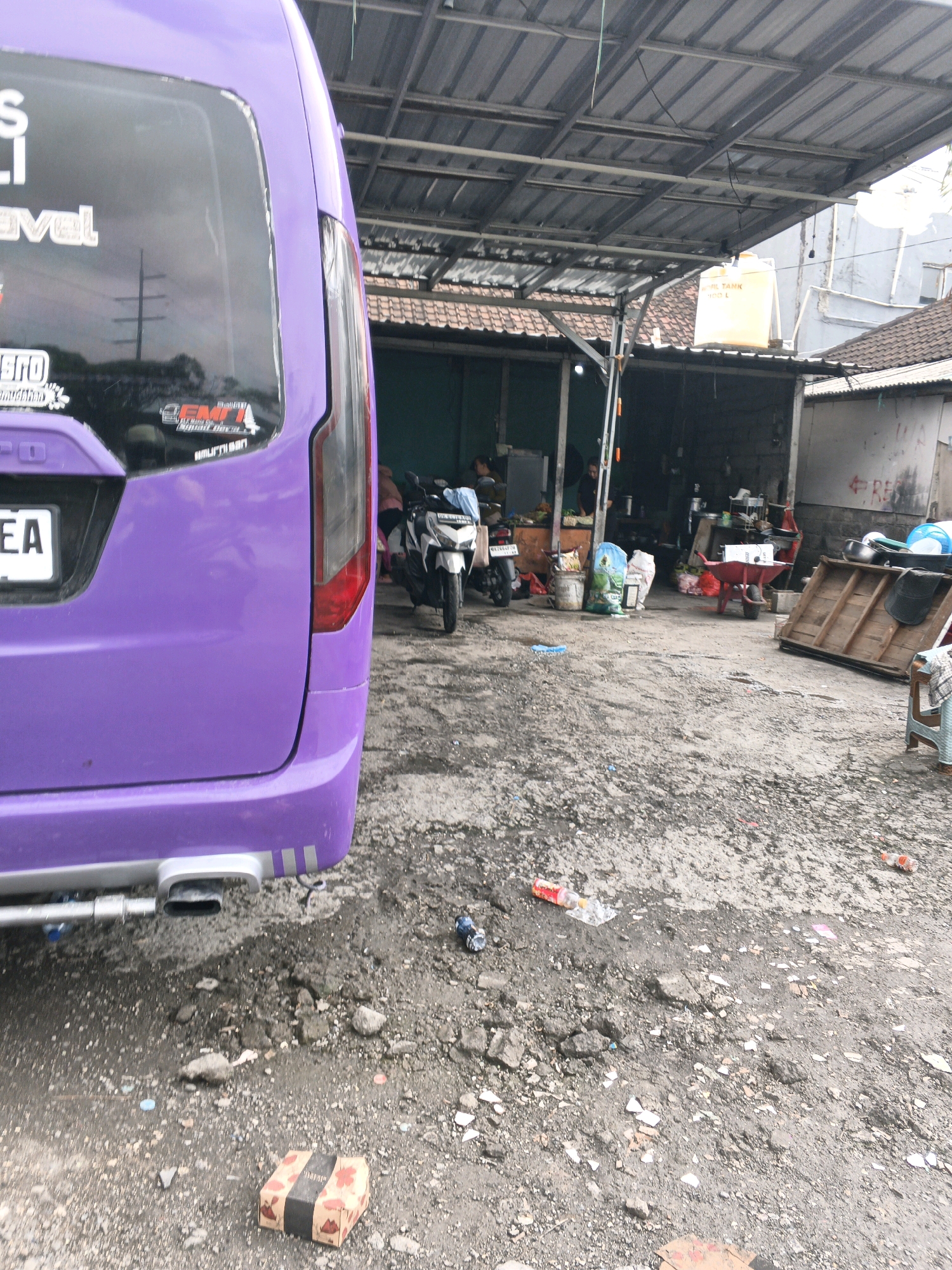 A purple minivan is parked at a roadside establishment in Denpasar, Indonesia. The van's rear is facing the camera, with its license plate reading "EA". The vehicle is situated on a gravel and dirt ground, surrounded by various items. To the left of the van, a metal exhaust pipe is visible. In the background, a covered area with a corrugated metal roof is set up, likely a food stall or repair shop. Several motorcycles are parked under the shelter, and there are people in the background. A yellow water tank is perched on the roof of a building behind the covered area. The ground is littered with debris, including plastic bottles and wrappers. A red wheelbarrow is also present, along with wooden planks. The overall scene depicts a bustling, outdoor commercial area with a focus on vehicle services or local trade.
