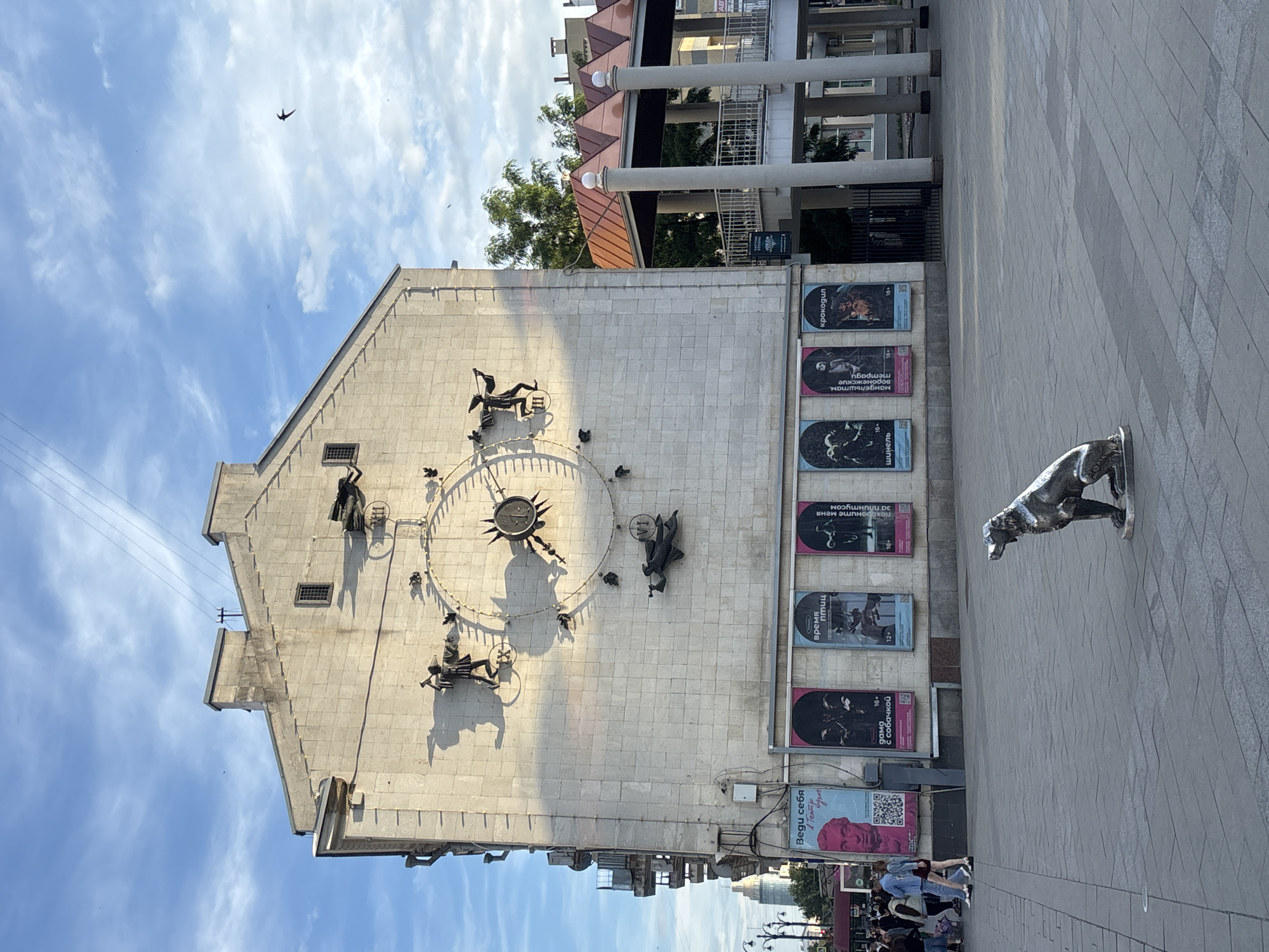 This is a vertical, full-shot photograph of an outdoor scene in Novaya Usman', Russia. The image is dominated by a large, light-colored building with a unique clock face featuring several sculptural figures. The clock face itself is circular with Roman numerals and a sun-like central element. Surrounding the clock are various metal figures, some appearing to be people in dynamic poses.
To the right of the building, a paved walkway is visible, leading to a sculpted metal dog positioned in a walking stance. The dog is the most prominent object in the foreground. Along the building's facade, several posters are displayed vertically. These posters contain text in Russian, with some indicating age restrictions like "16+" and "12+". Some of the readable text includes "Крокодил" (Crocodile), "время птиц" (time of birds), and "дама с собачкой" (lady with a dog).
The sky above is a bright blue with scattered white clouds. A single bird is captured in flight against the sky, adding a dynamic element to the scene. The overall lighting suggests it is daytime, possibly late morning or afternoon, given the shadows cast by the figures and the building. The presence of people on the walkway in the lower right portion of the image indicates it's a public area. The scene appears to be a town square or a pedestrian area in front of a building, possibly a cultural center or theater, given the posters. The mood is neutral, depicting a typical urban environment.